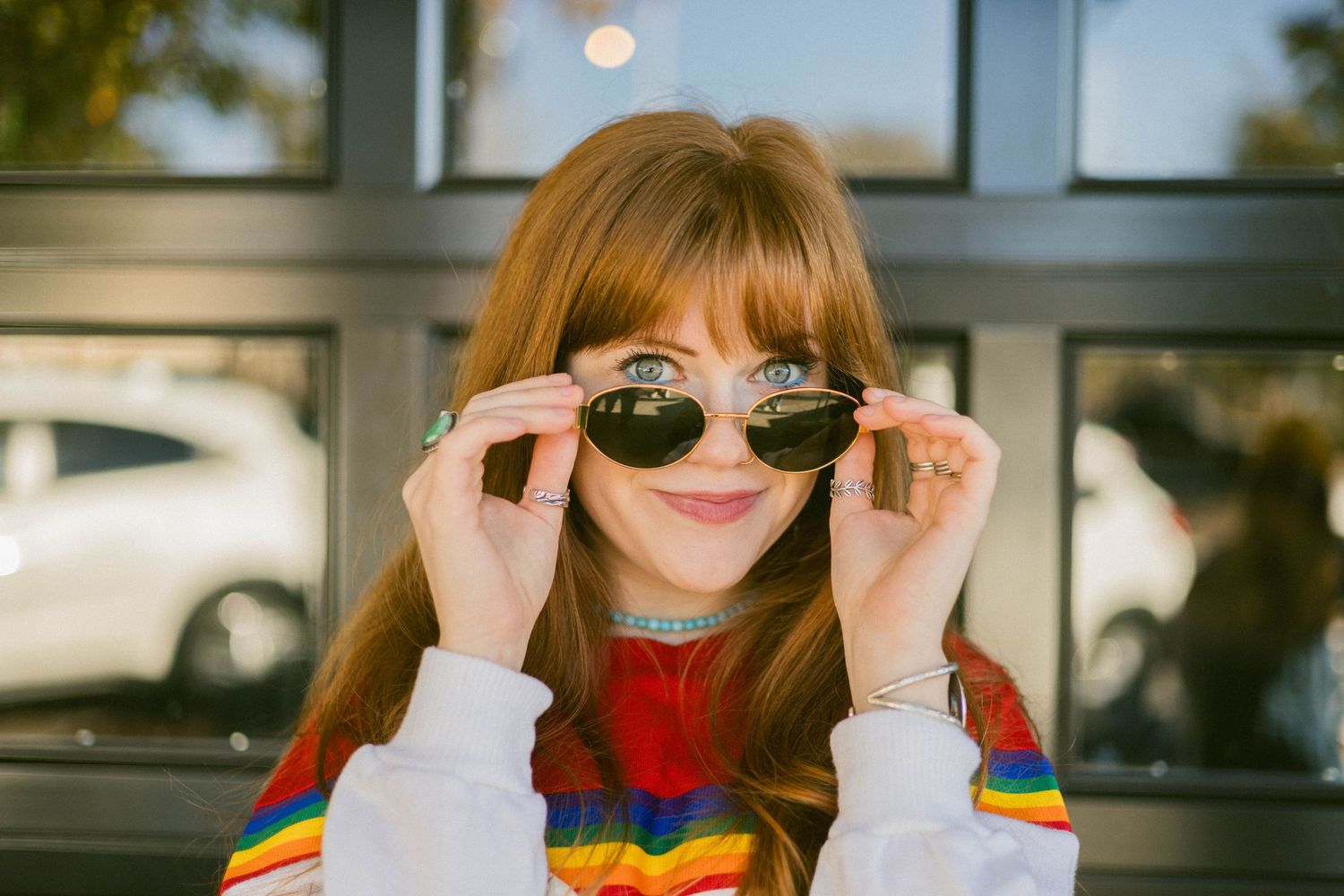 Person in a colorful striped sweater poses playfully with sunglasses against a bright outdoor background.