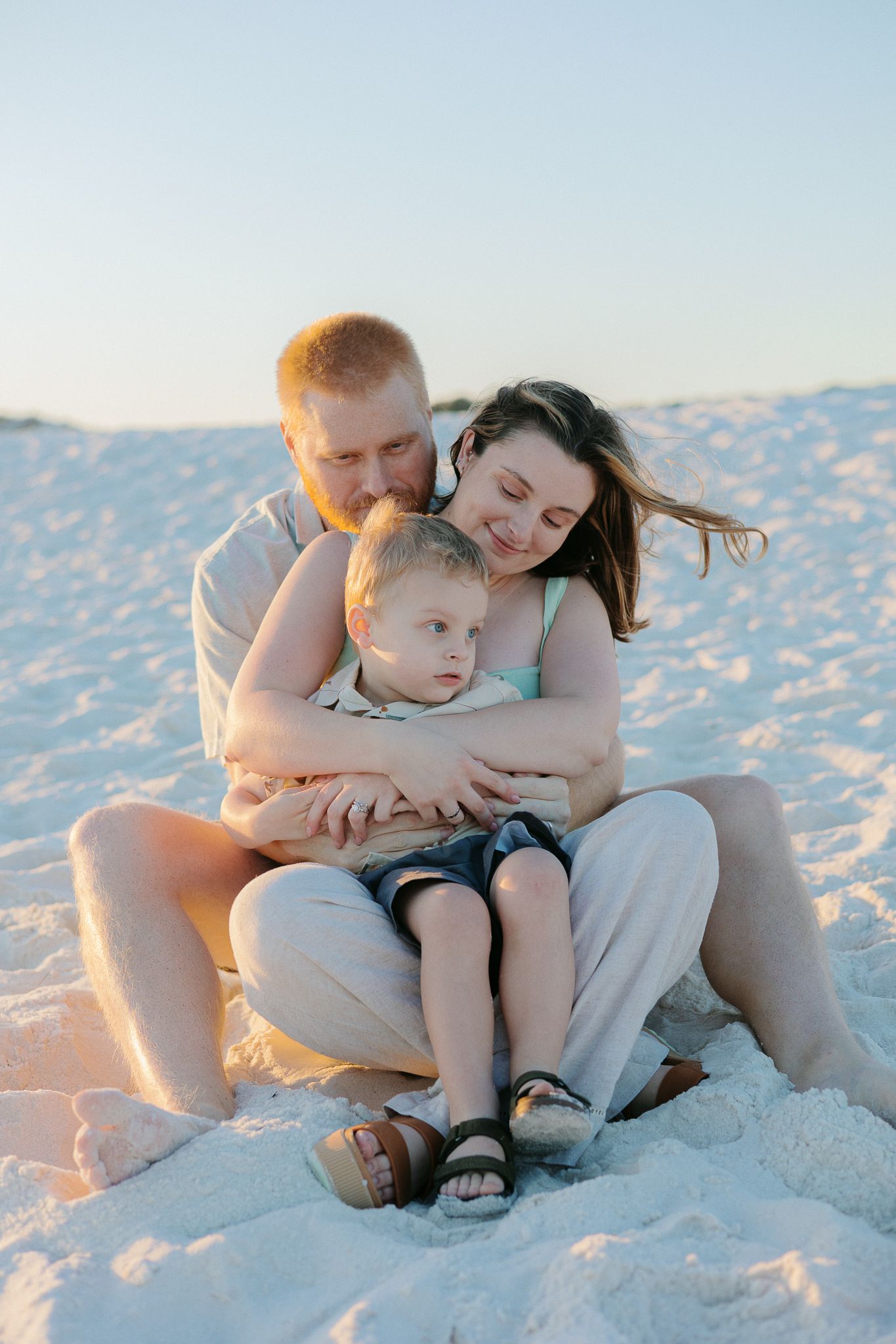 Family of three sitting on white sand beach at sunset, parents embracing young son with clear sky backdrop.