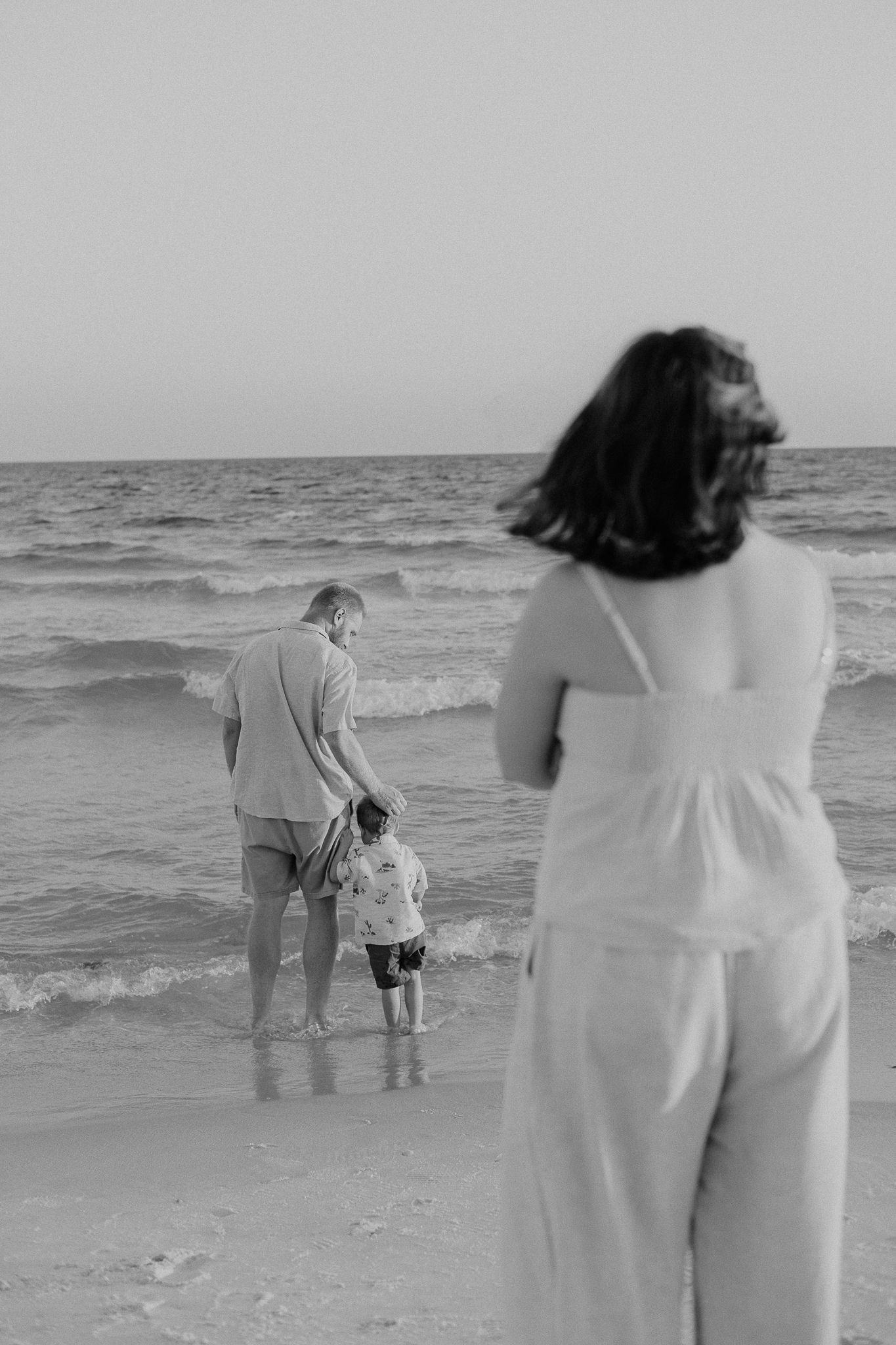 Black and white photo of a family at the beach, father and child standing at the water's edge while mother watches.