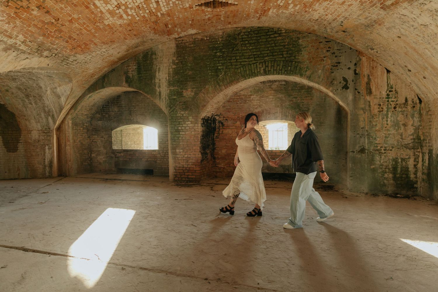 Two people in flowing outfits dance in dramatic underground brick tunnel with natural light.