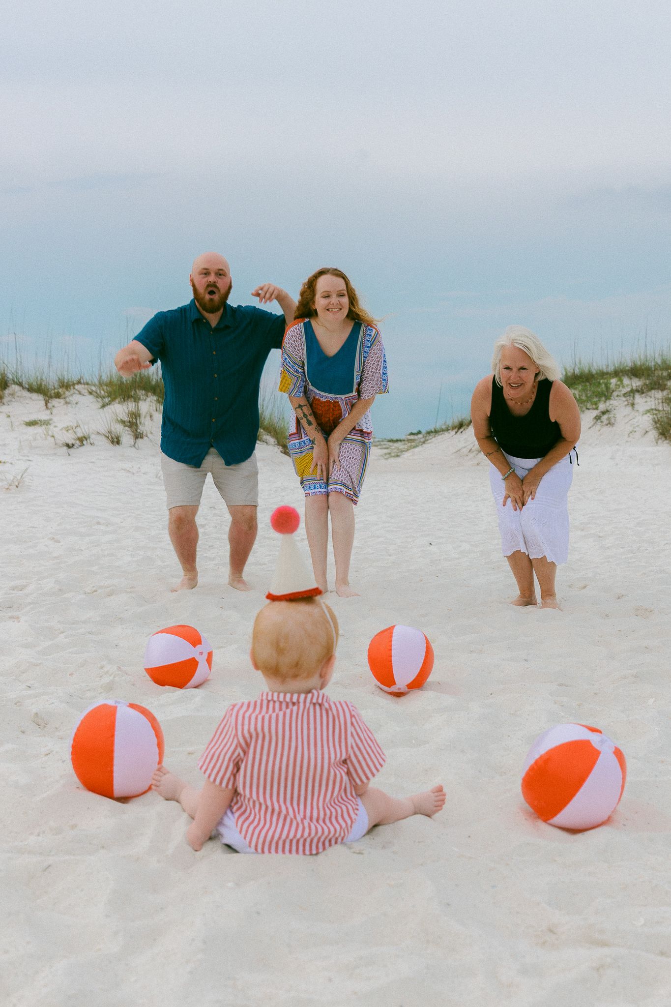 Family playing with colorful beach balls on white sand beach with baby sitting in foreground near sand dunes.