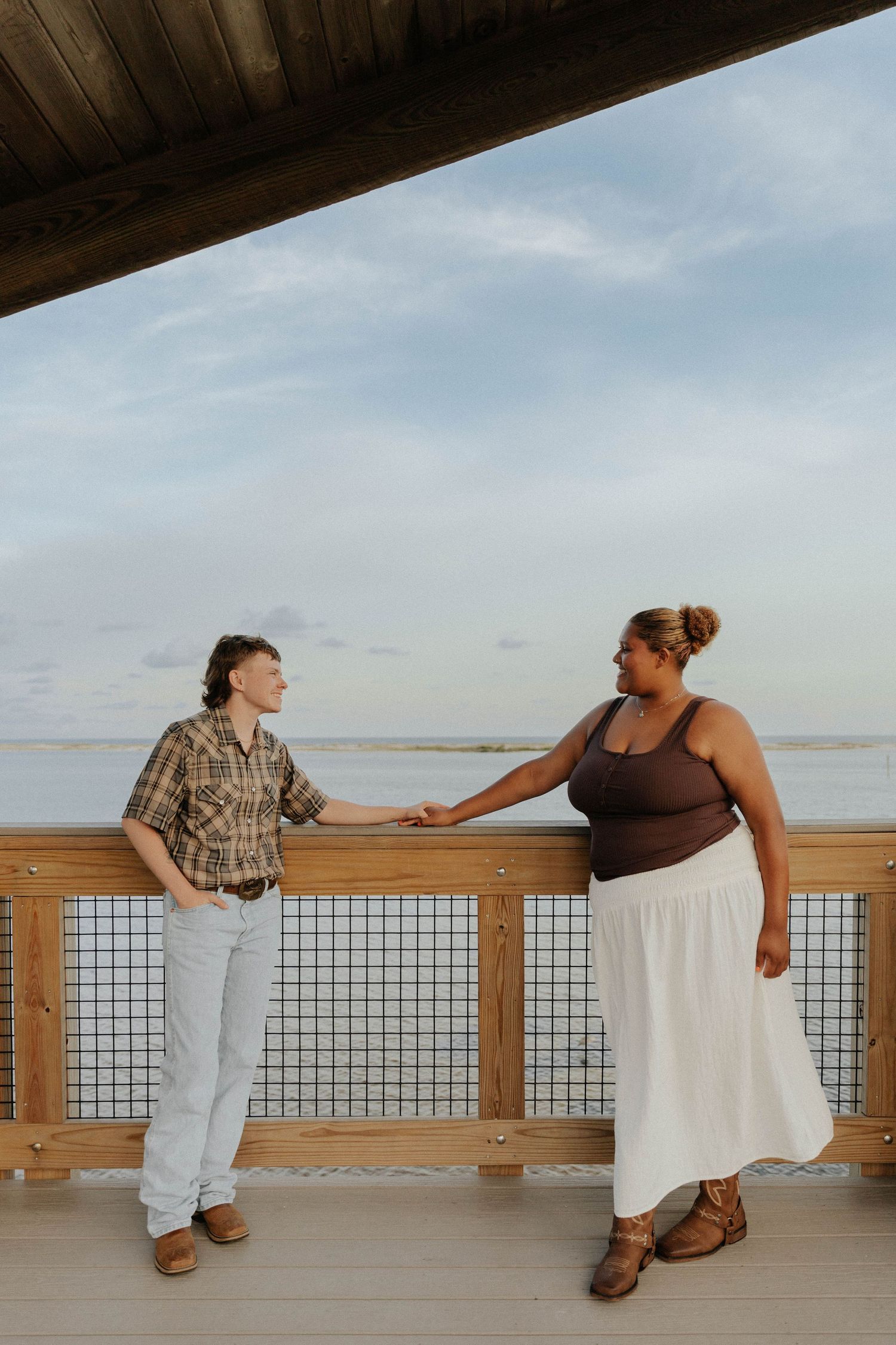 Two people lean on a wooden railing overlooking the ocean in a series of playful poses at sunset.