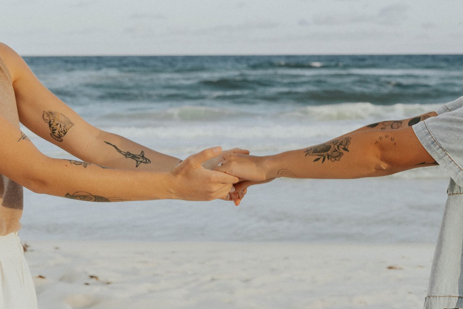 Two tattooed arms reach out to hold hands against an ocean backdrop.