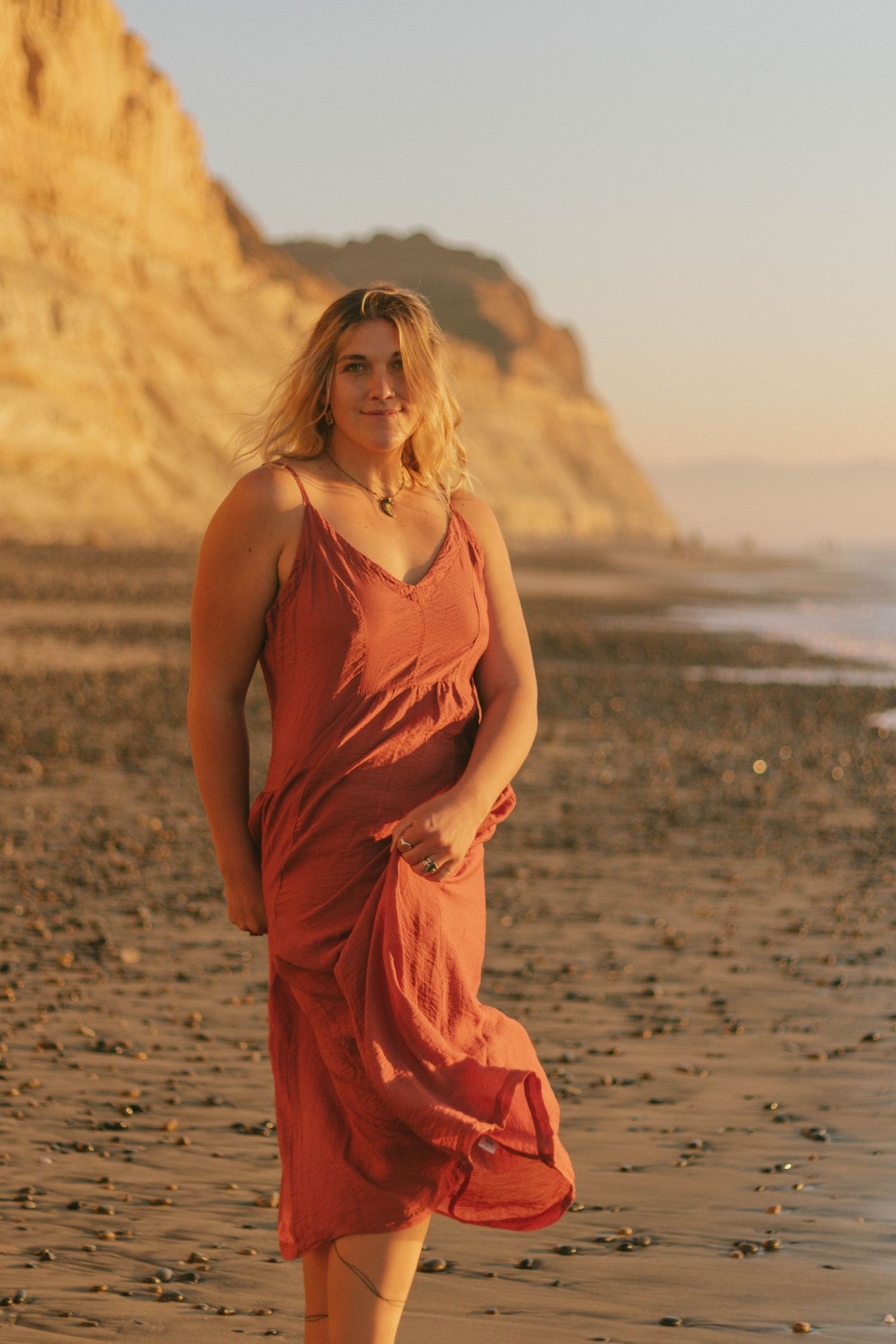 Person in flowing coral dress poses on sandy beach at sunset with cliffs in background.