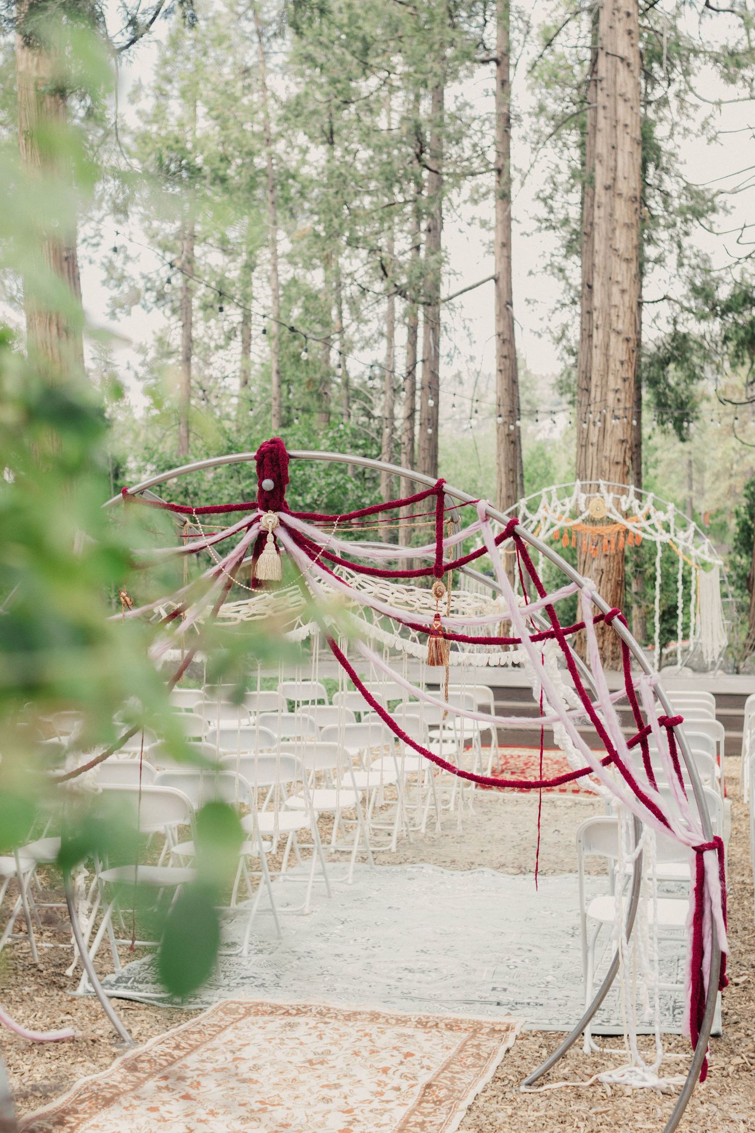 Outdoor wedding ceremony setup with burgundy draped fabric arch and white chairs in a forest setting.