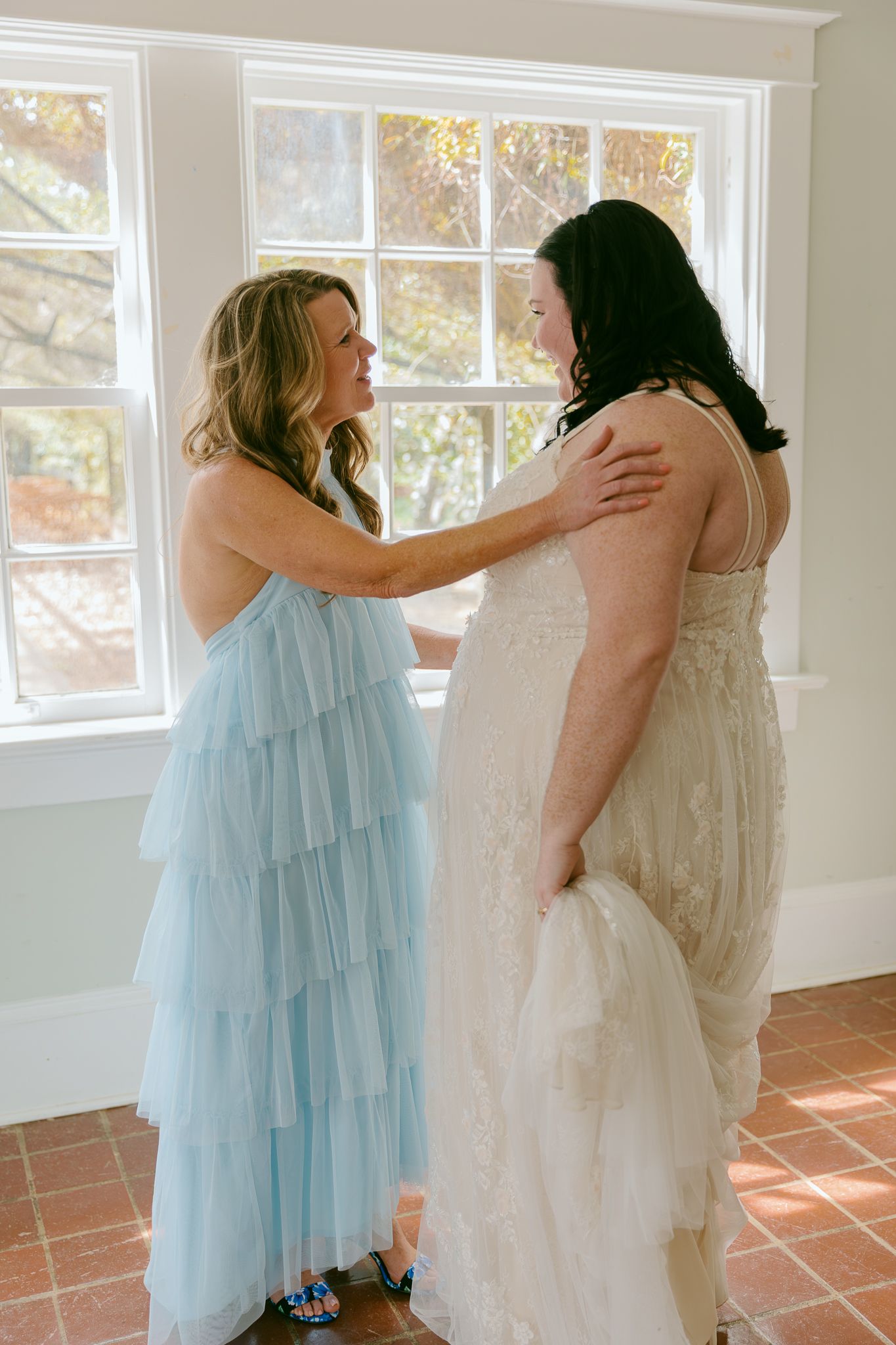 A bridesmaid in a light blue tiered dress helps a bride in a white lace gown prepare near a sunlit window.