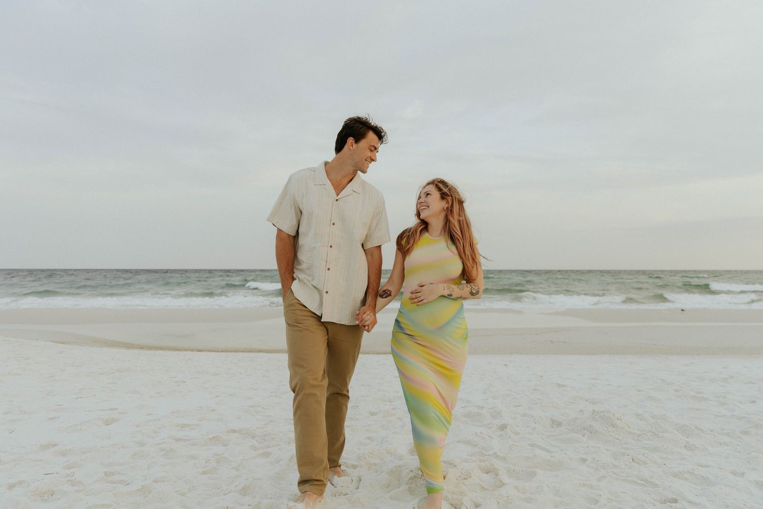 A couple walks together on a beach at sunset while wearing casual attire and sharing a joyful moment.