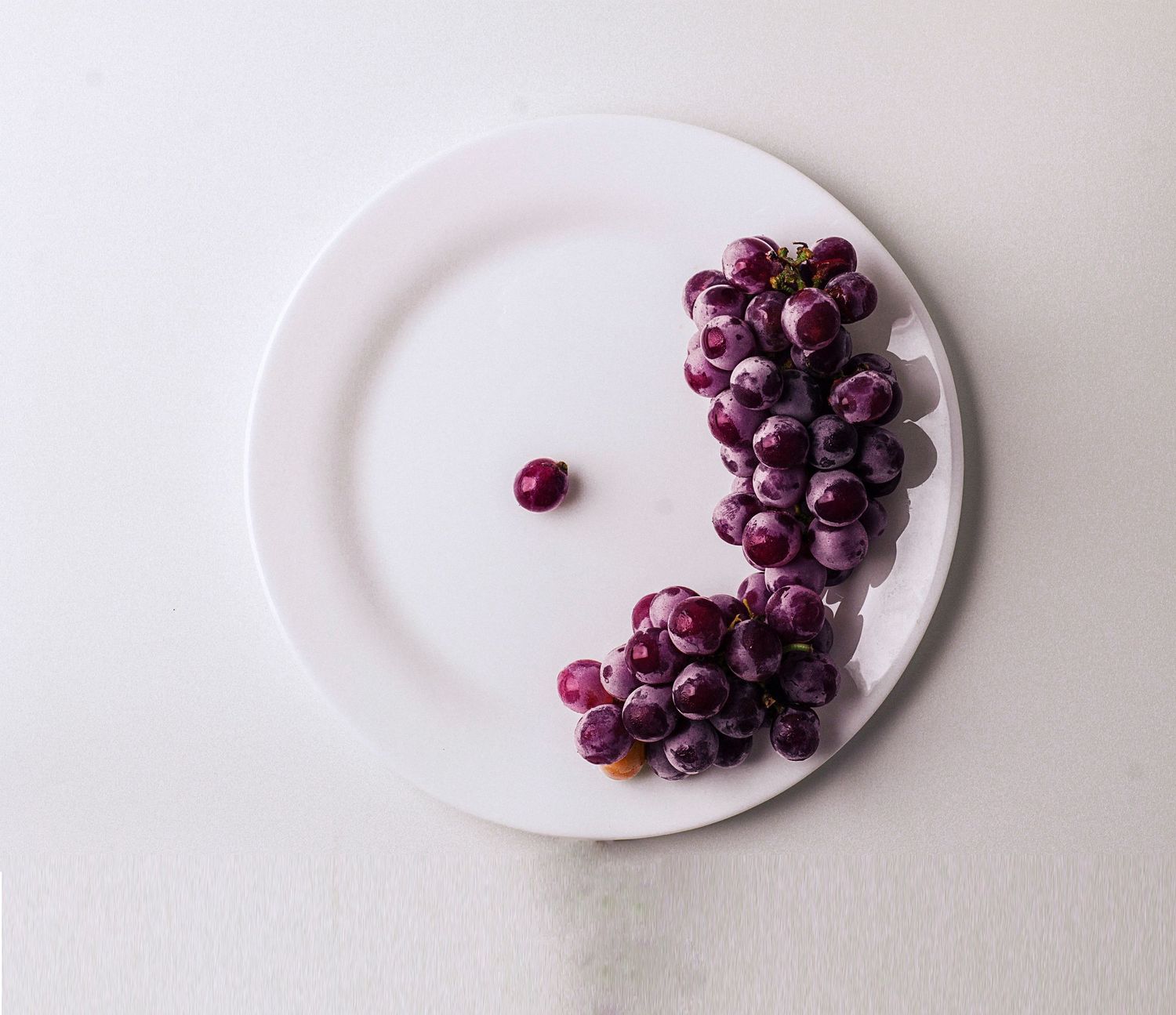 Cacho de uvas roxas com uma única uva solta ao centro de um prato branco. Minimalismo visual com frescor e elegância.