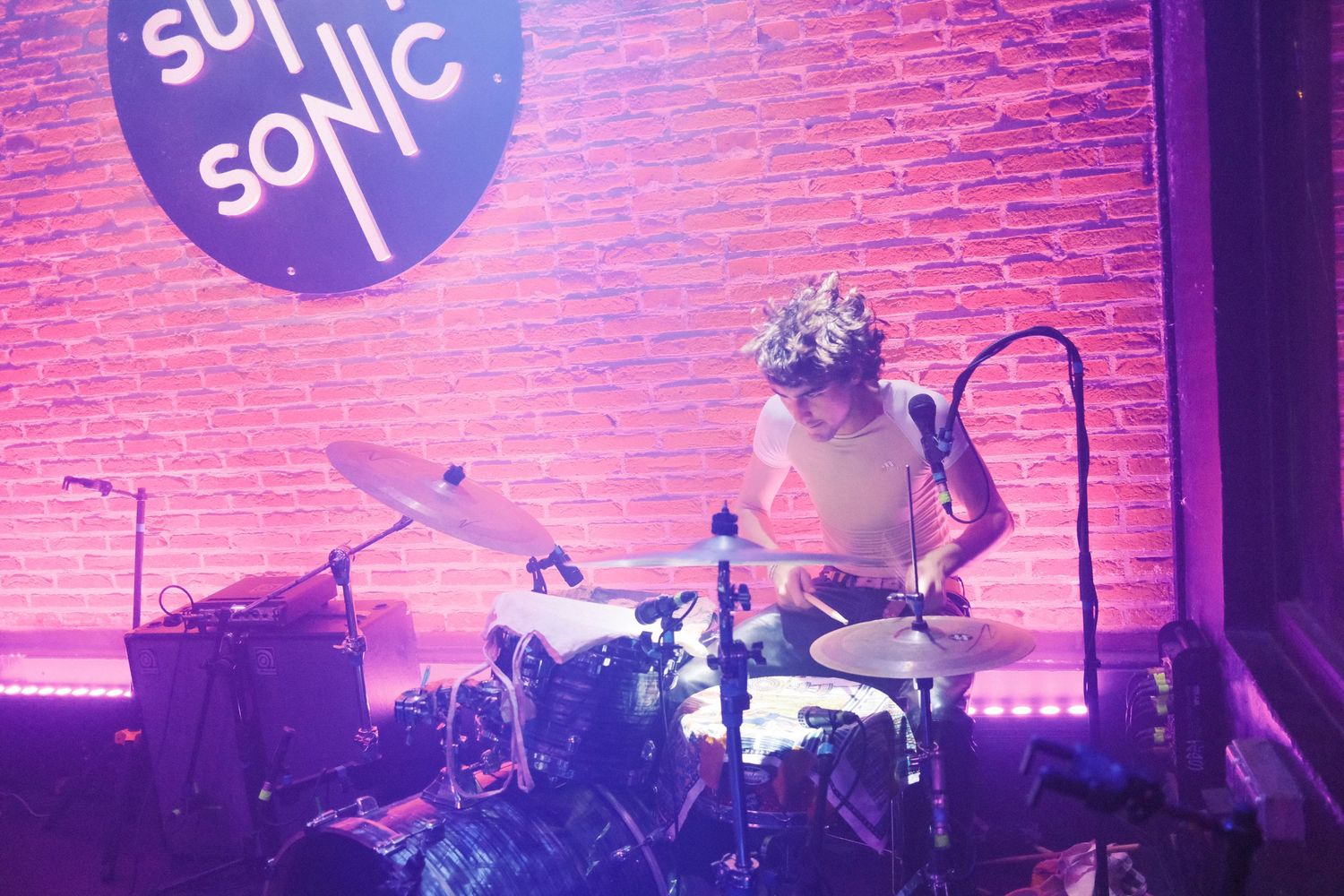 un batteur au supersonic sur des lumières violettes joue de la batterie, concentré il ne regarde pas devant lui