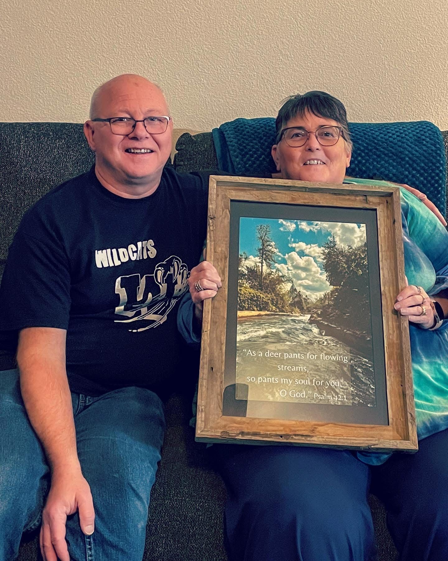 Example of a past order. Couple smiling on couch holding a rustic framed Psalm 42:1 scripture framed print with forest stream photo.
