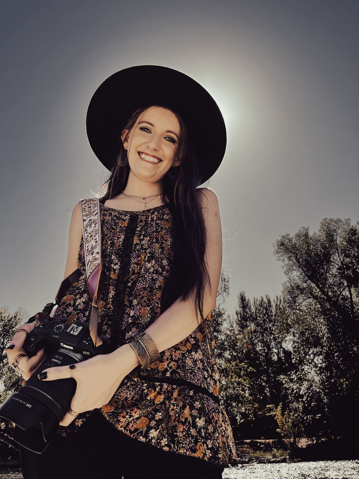 Owner, creator and designer of Farm & Folklore Smiling woman in black wide-brim hat holds camera outdoors with sun halo behind her.