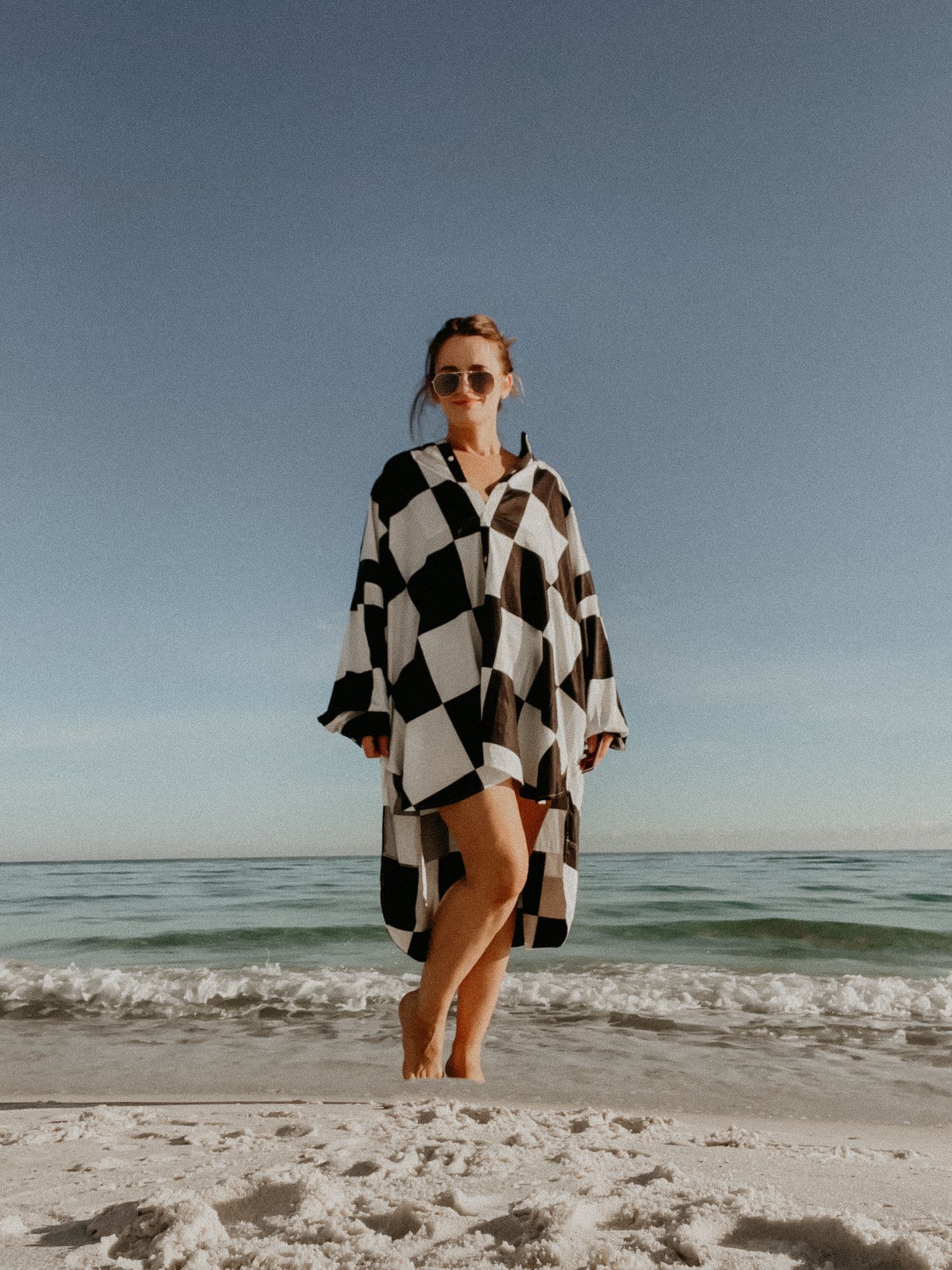 Woman in black and white checkered dress walking barefoot on a sandy beach with ocean waves behind her.