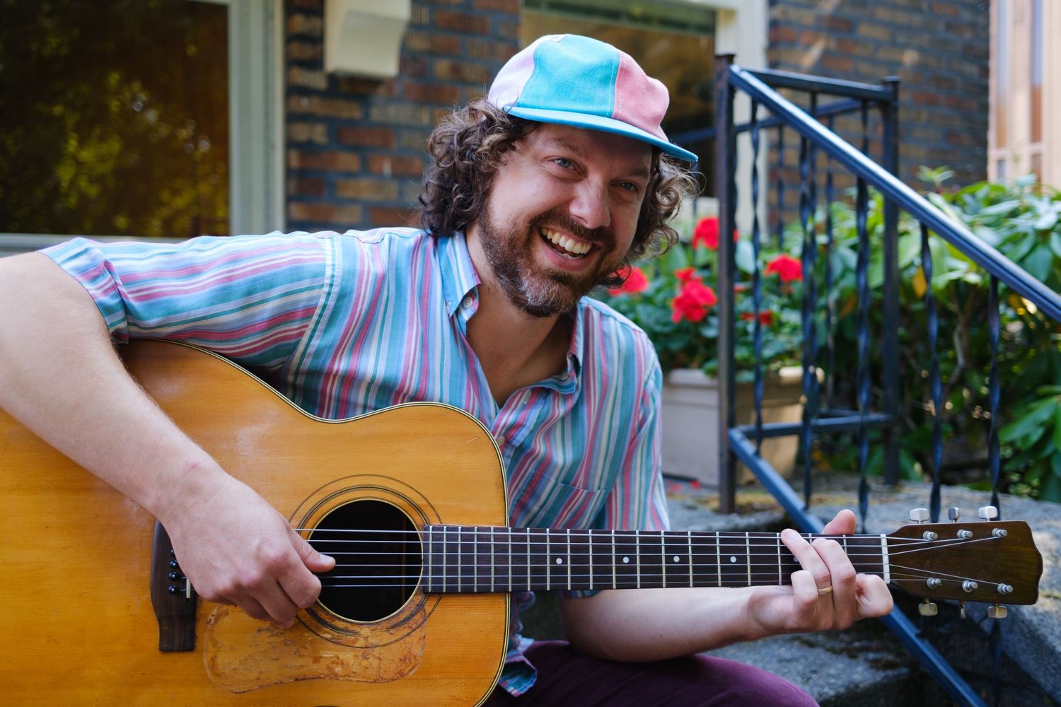A musician plays acoustic guitar while sitting outdoors near brick buildings.