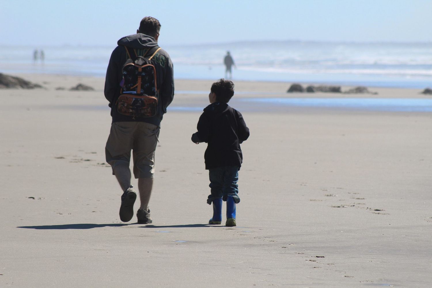 Adult and child walking hand in hand along a sandy beach shoreline, with ocean waves and blue sky in the background.