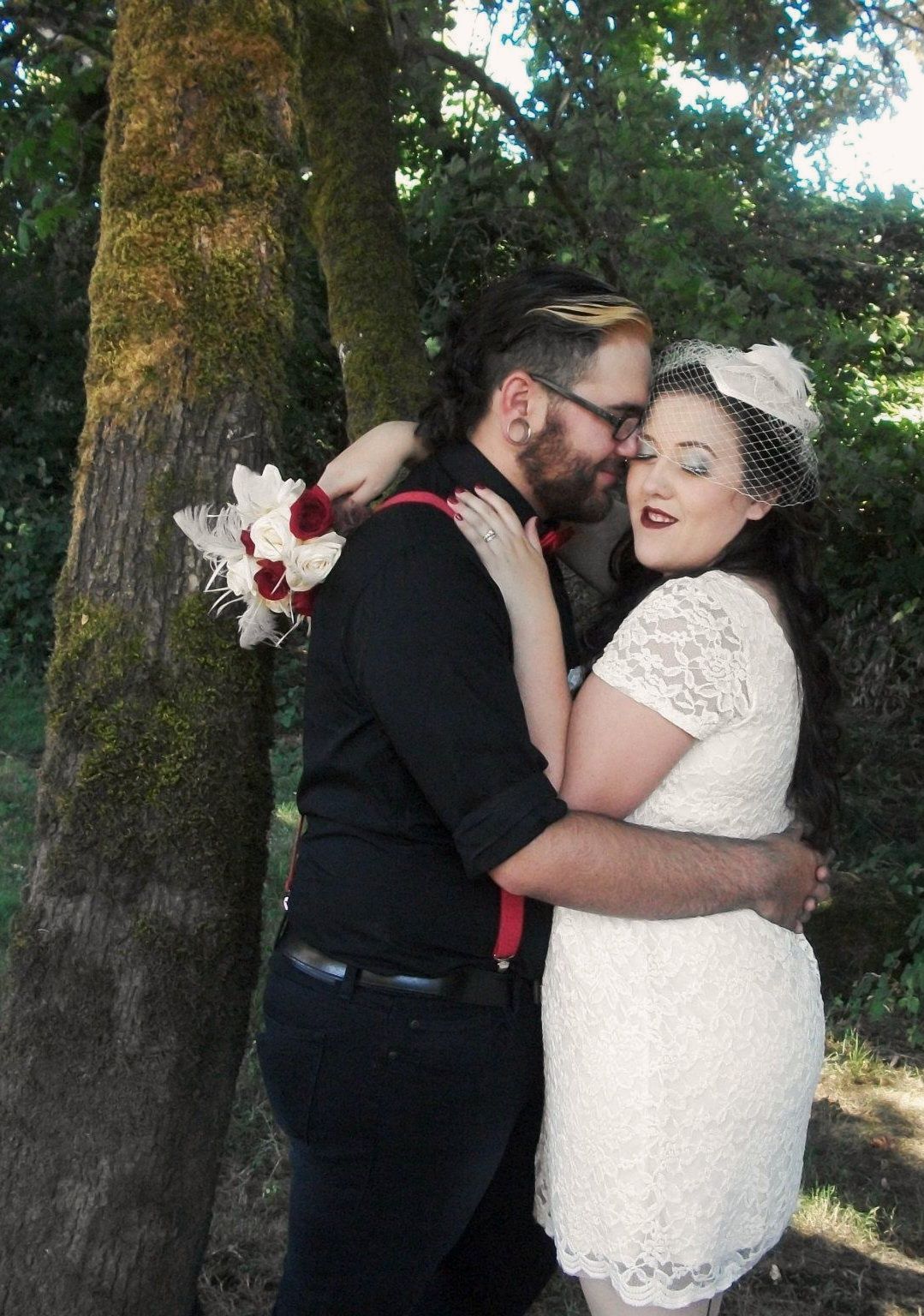 Bride and groom embrace near a mossy tree, she wears lace dress and birdcage veil, he wears black shirt with red suspenders.