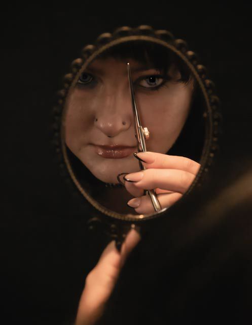 Woman gazing at reflection in ornate vintage hand mirror while holding decorative scissors near her face to cut her hair hairstylist photo