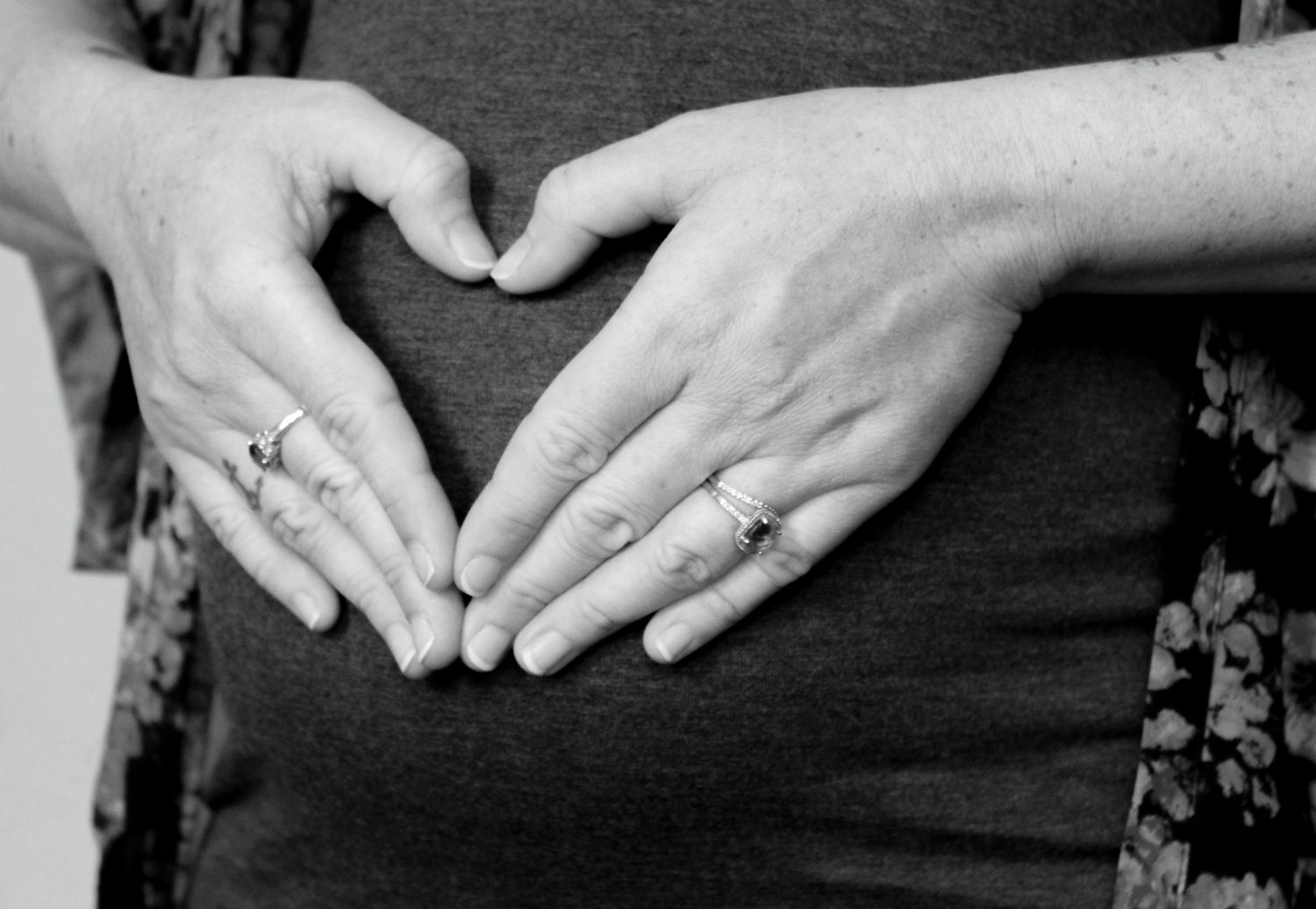 Close-up of woman forming heart shape with hands over pregnant belly in black and white.