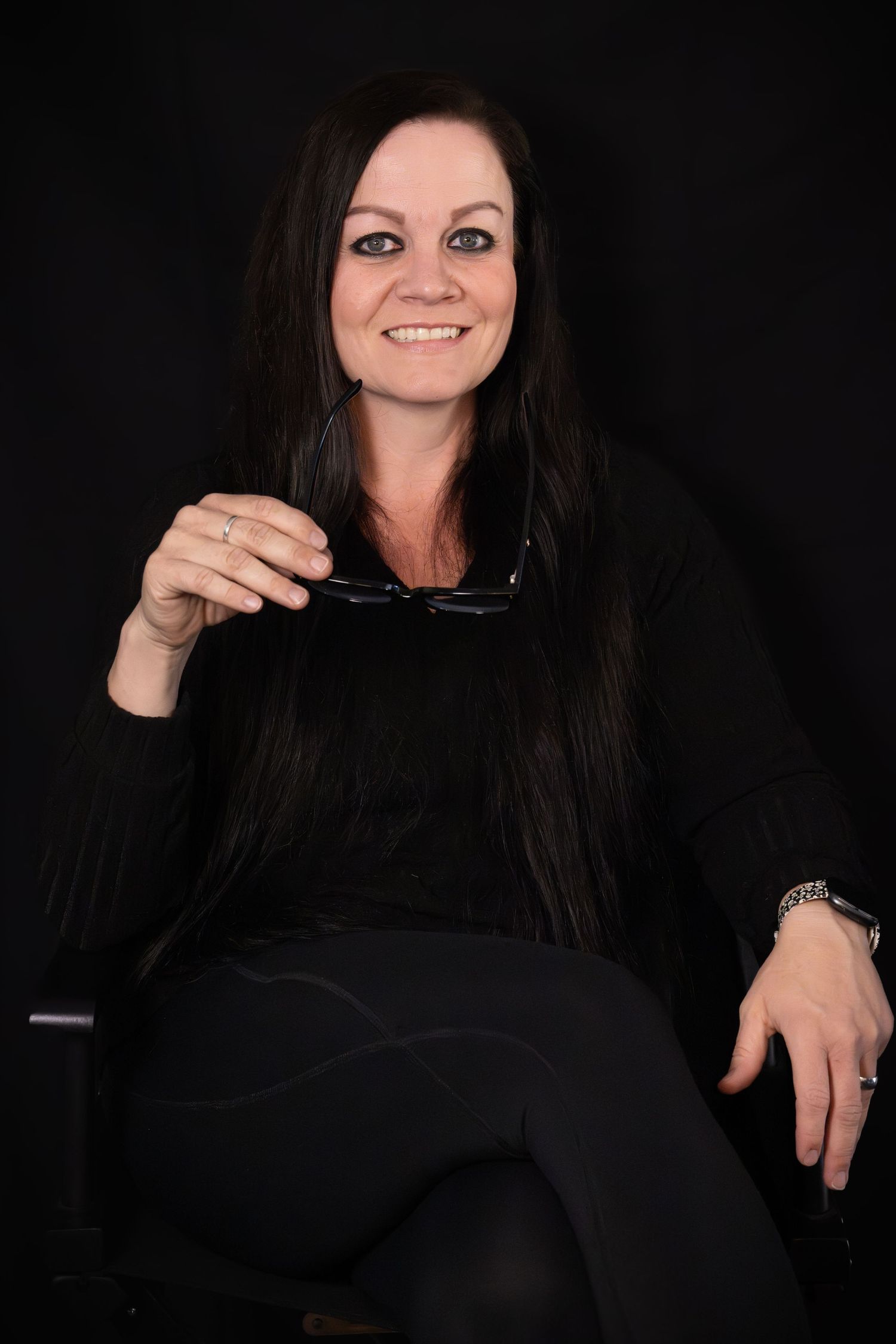 Smiling woman with long dark hair sitting in chair holding glasses, wearing all black outfit against dark background.