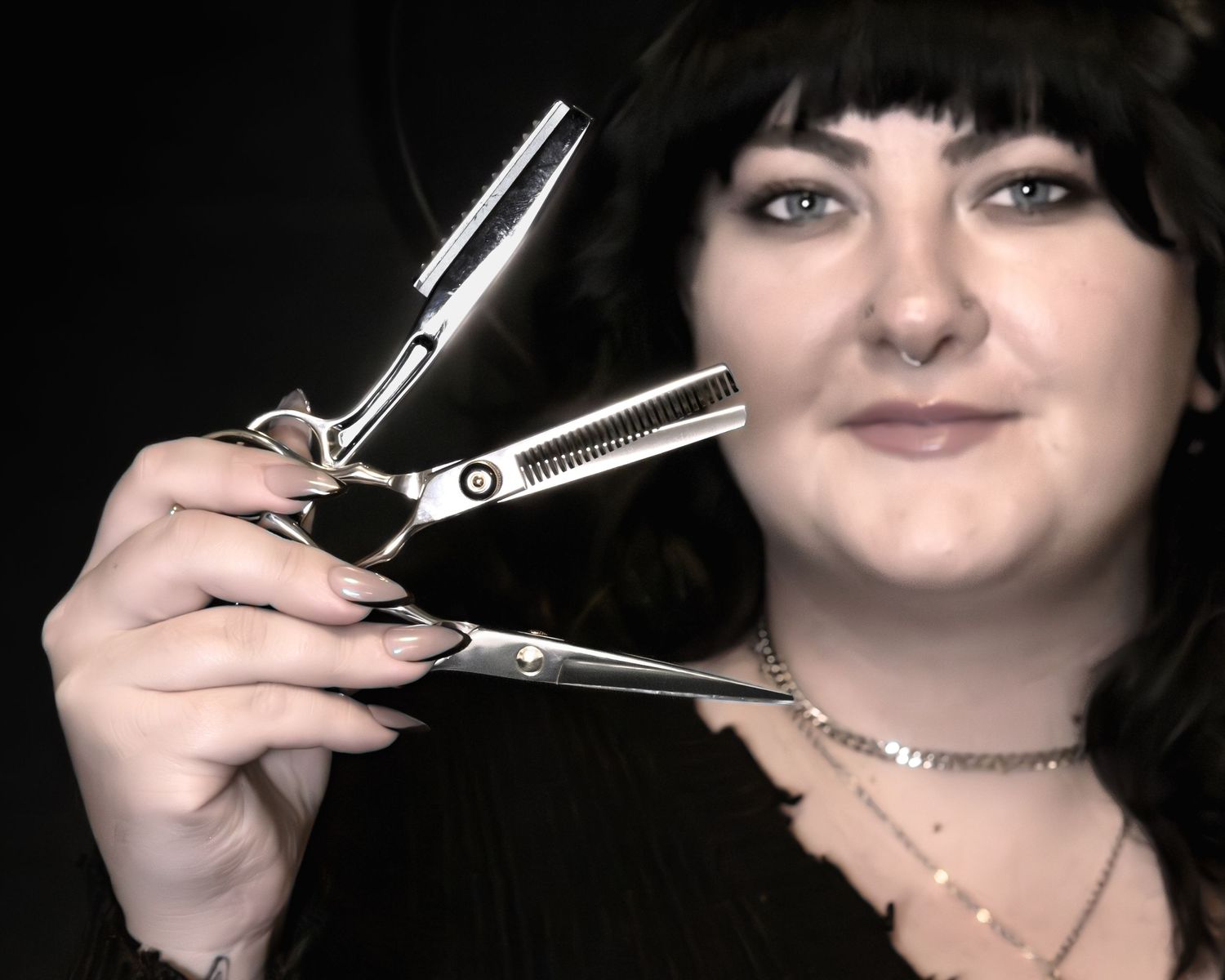 A smiling female hairstylist holds professional thinning scissors and a razor comb, showcasing her nail art in a dark studio.
