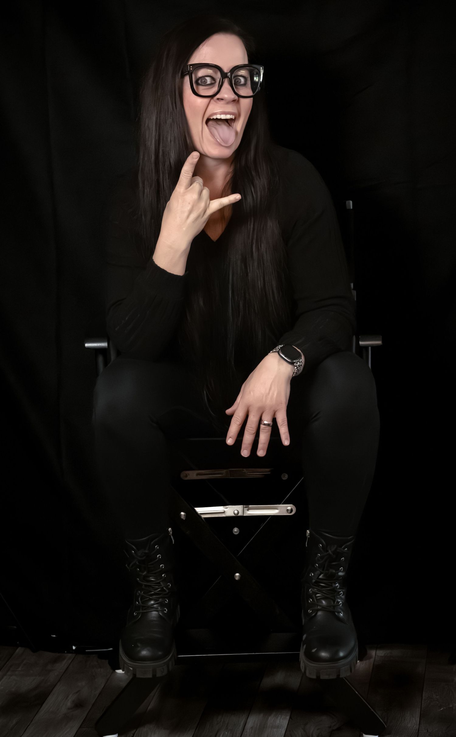 Woman in black outfit and glasses makes rock sign gesture while sitting in chair against dark background.