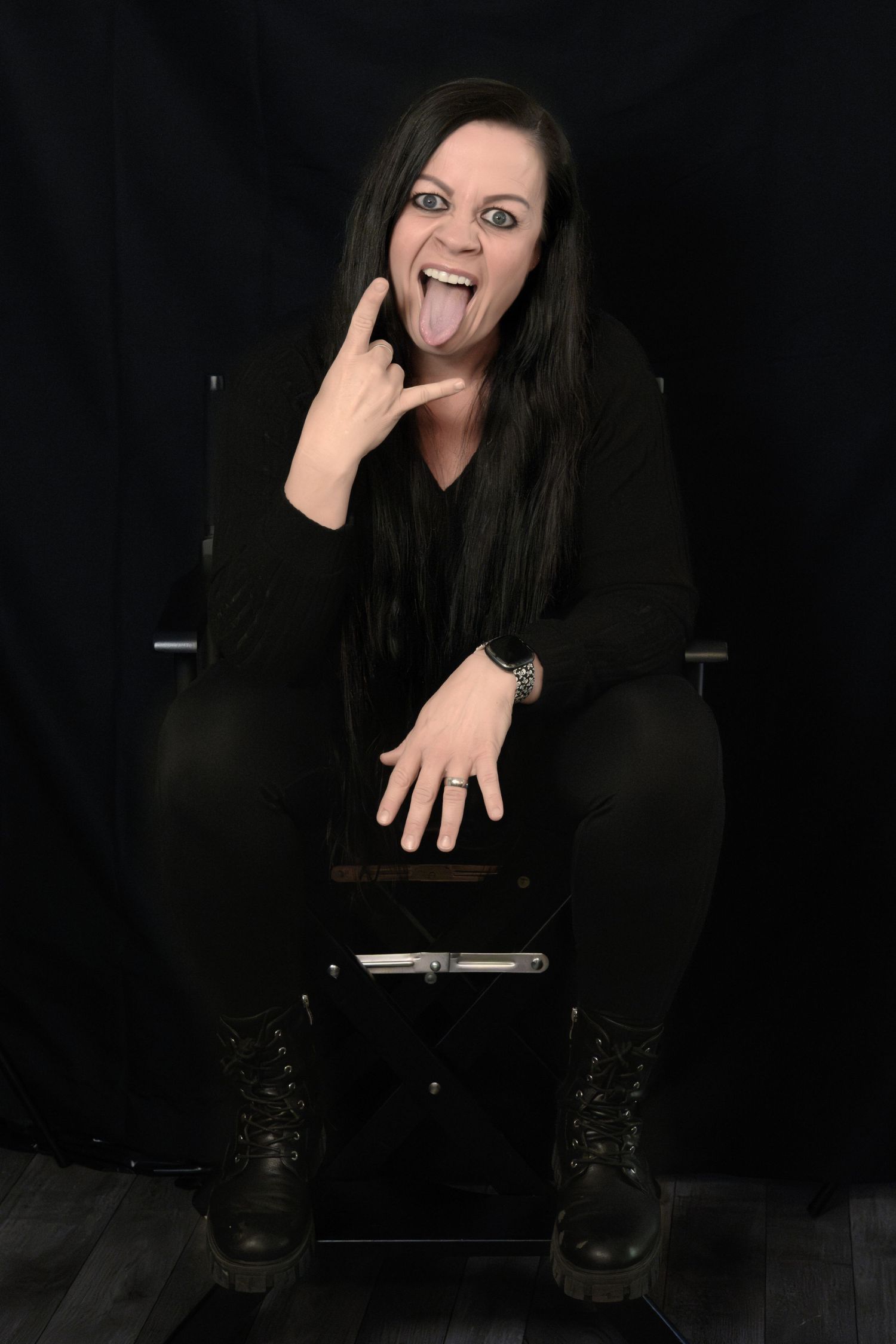 Woman in black makes rock sign with tongue out, seated on stool against dark backdrop.