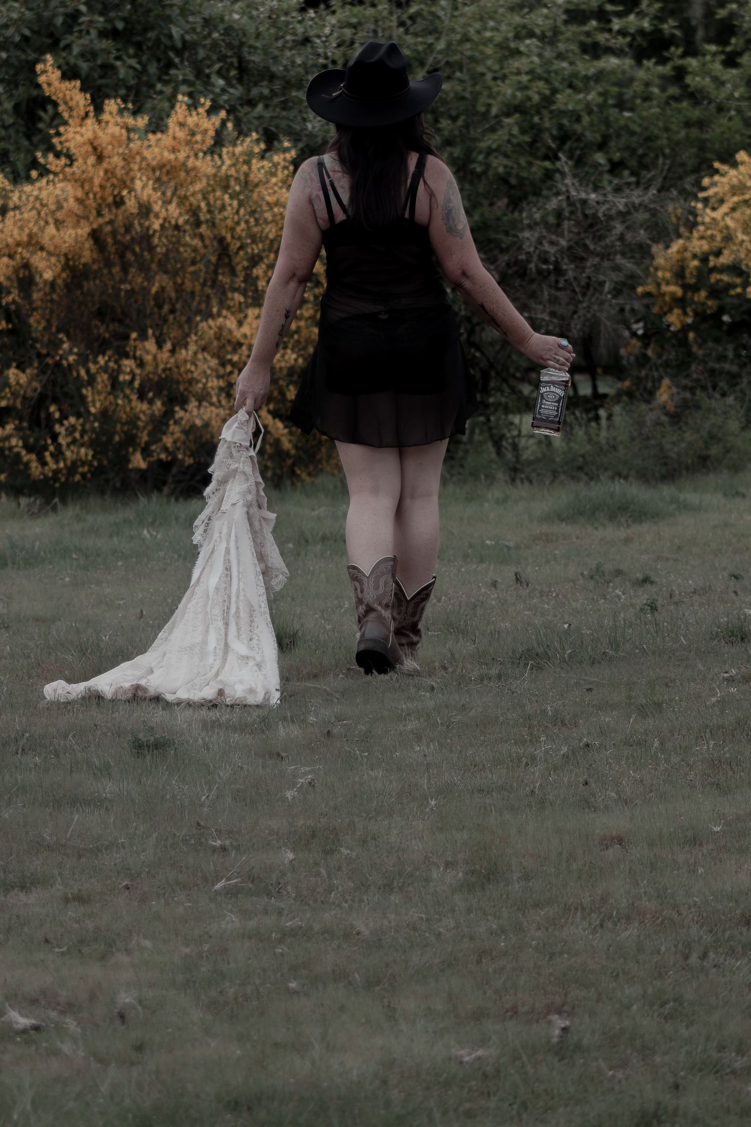 Woman in black dress cowboy boots and hat walking in field holding white dress and bottle.