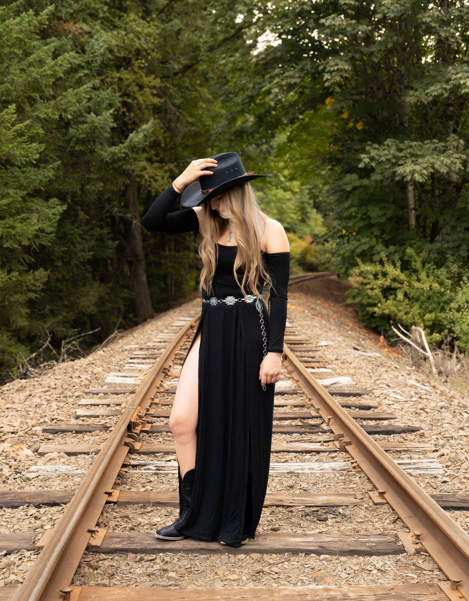 Woman in black off-shoulder gown, wide-brim hat, and boots posing on railroad tracks surrounded by lush green trees.