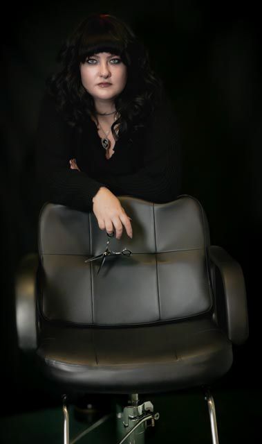 A female hairstylist in black poses behind a salon chair holding scissors, with dark curly hair and dramatic makeup.