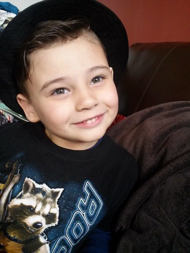 A smiling young boy wearing a black hat and Guardians of the Galaxy shirt sits on a brown leather couch.