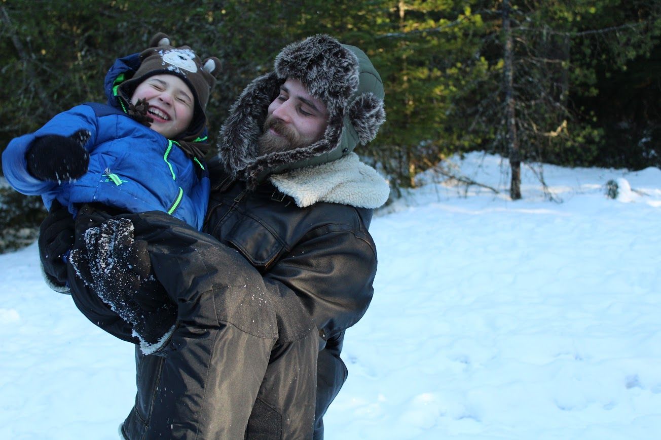 Man in fur hat holds laughing child in blue jacket while playing in snowy winter forest.