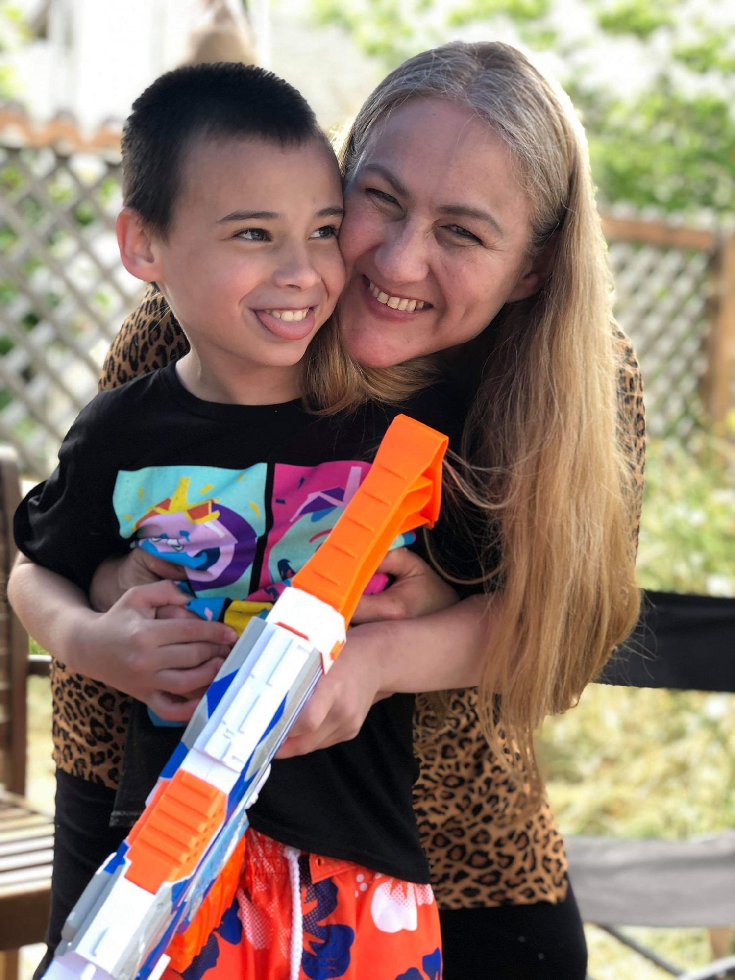 A woman with long blonde hair hugs a smiling boy holding a Nerf gun outdoors. Family, grandmother embraces grandson oregon