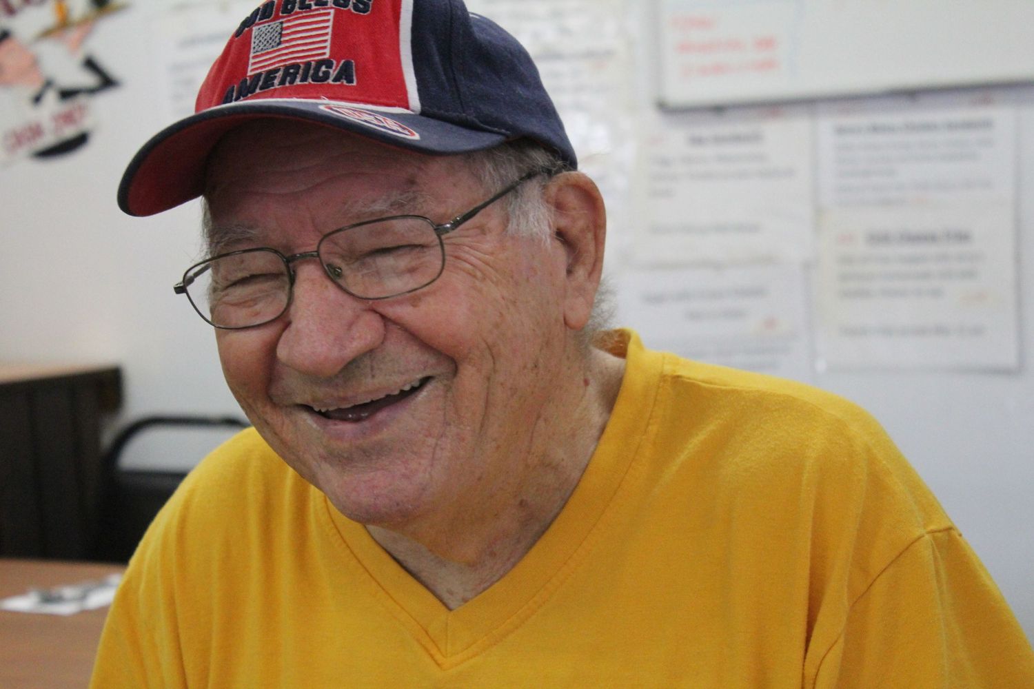 Elderly man smiling warmly, wearing a yellow t-shirt and a red America baseball cap, seated indoors. jerry's coffee shop simi valley CA