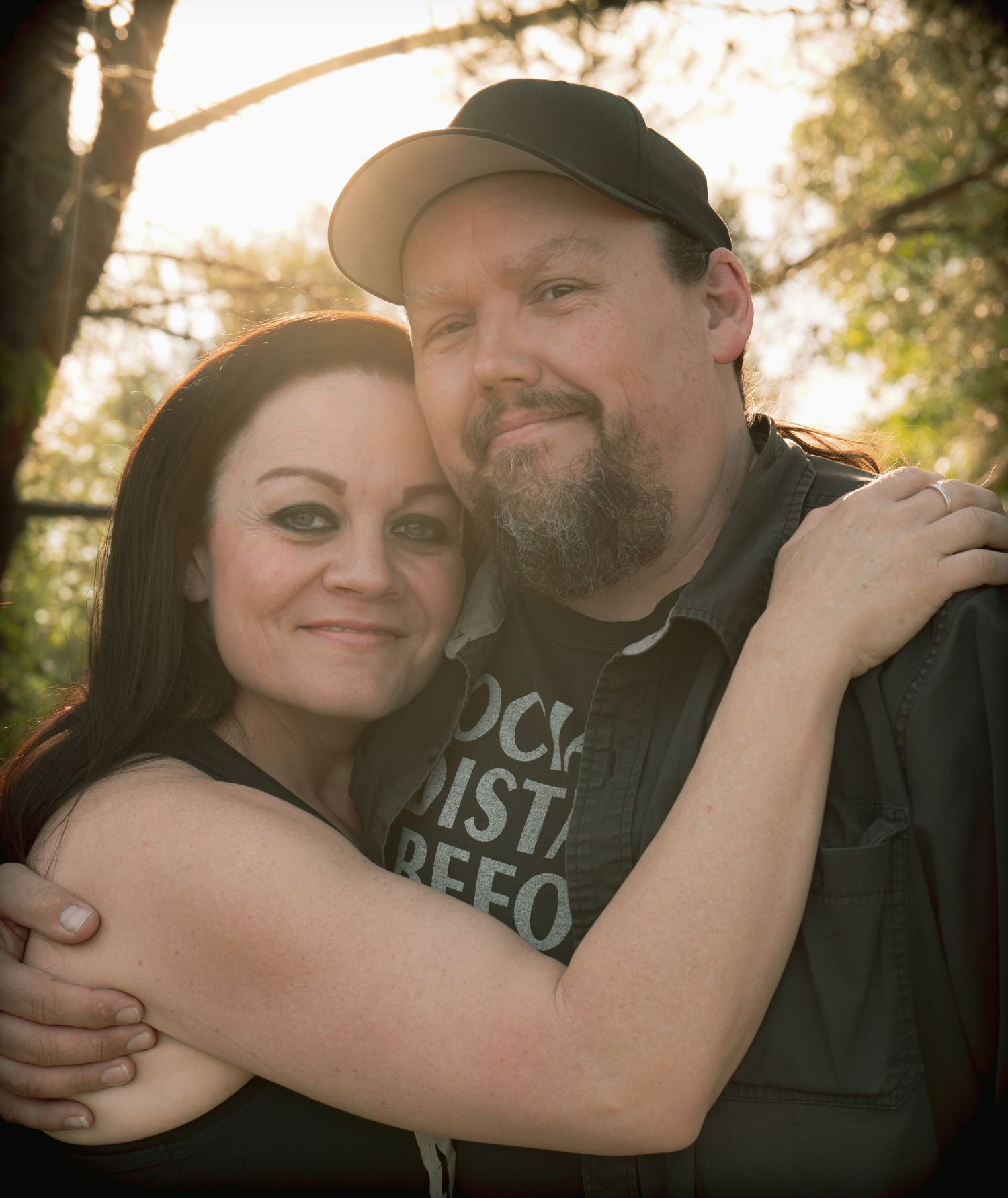 A couple embracing outdoors, smiling warmly, with trees and golden light in the background.