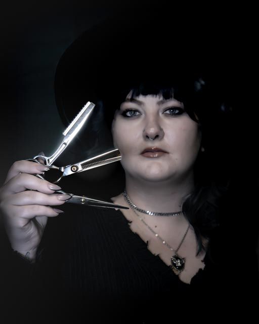 Gothic-styled woman in black hat holding straight razor and scissors in dramatic dark portrait.