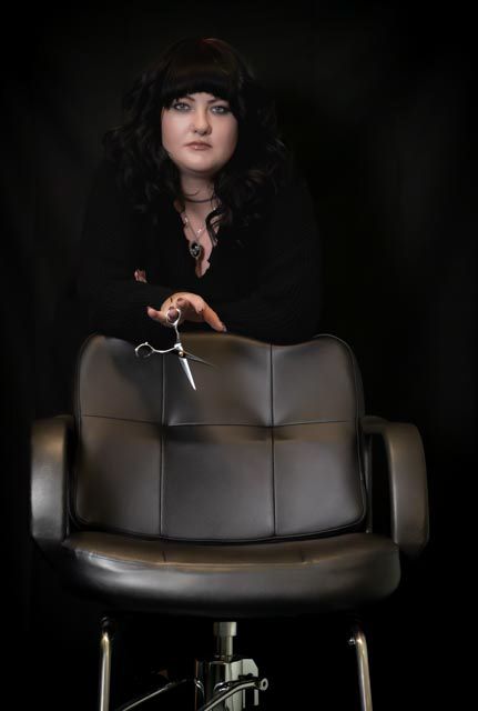 A female hairstylist in black clothing holds scissors over a salon chair against a dark background.