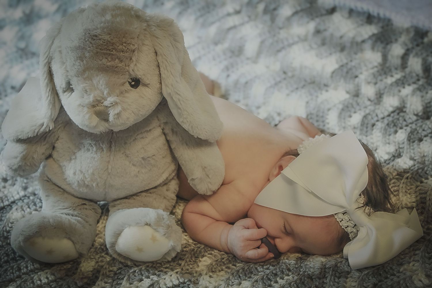 Newborn baby sleeping peacefully next to a soft gray bunny stuffed animal on a cozy blanket.