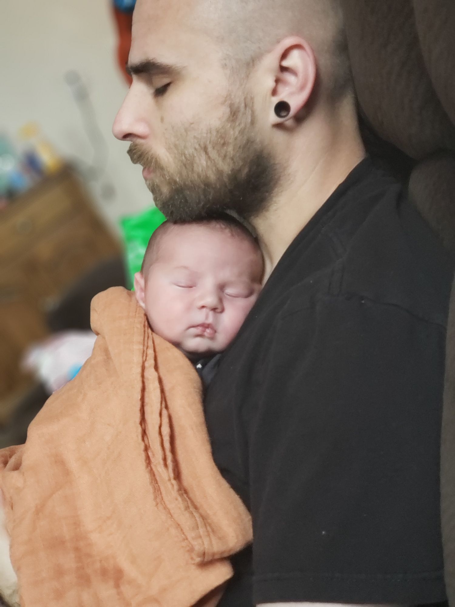 A bearded man with gauged ears holds a sleeping newborn baby wrapped in an orange blanket close to his chest.