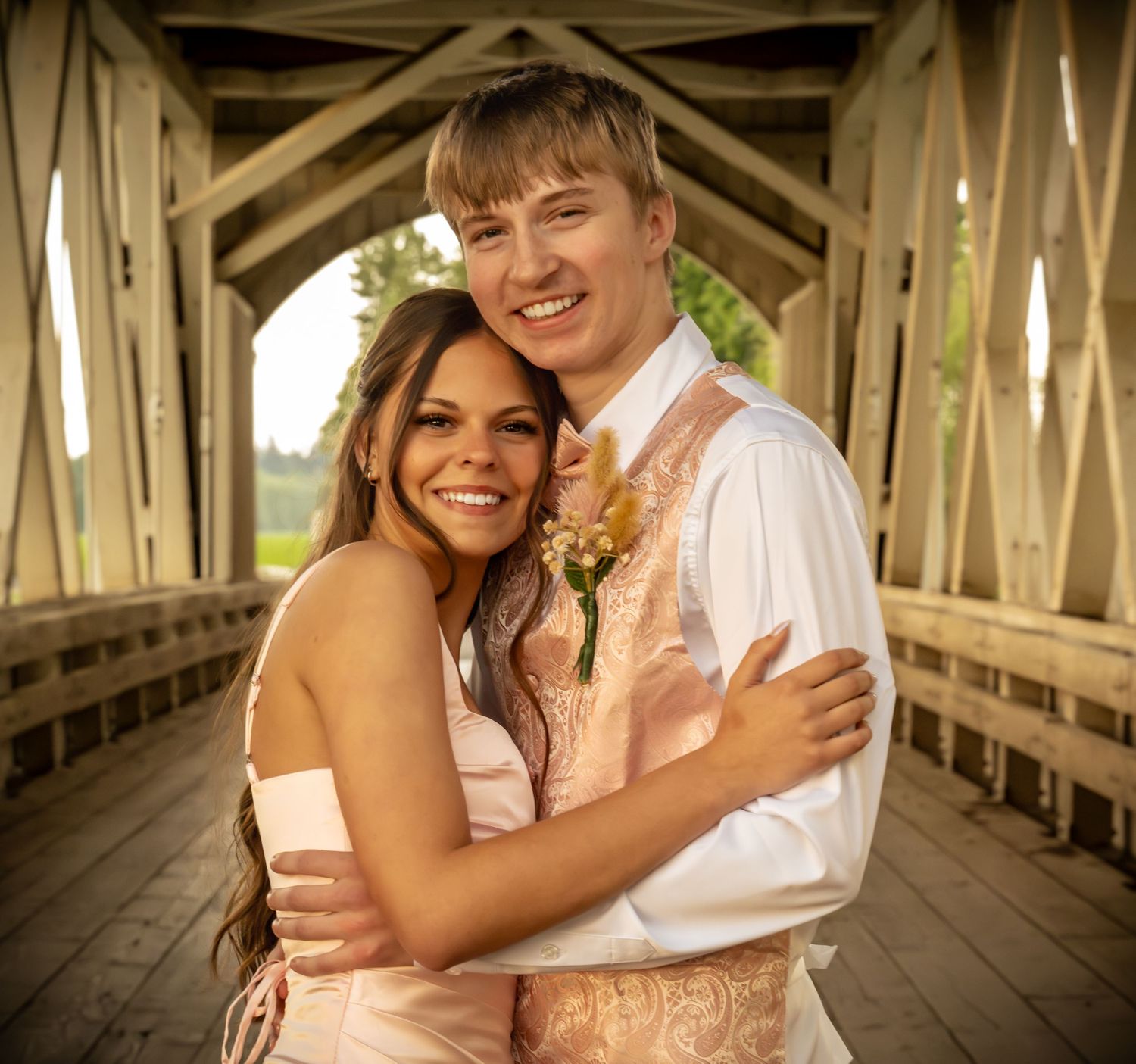 Young couple in formal attire embracing and smiling inside a rustic covered wooden bridge.