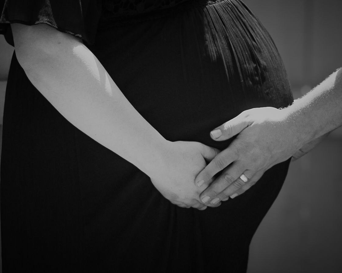 Black and white photo of couple holding hands over pregnant belly, man wearing wedding ring.