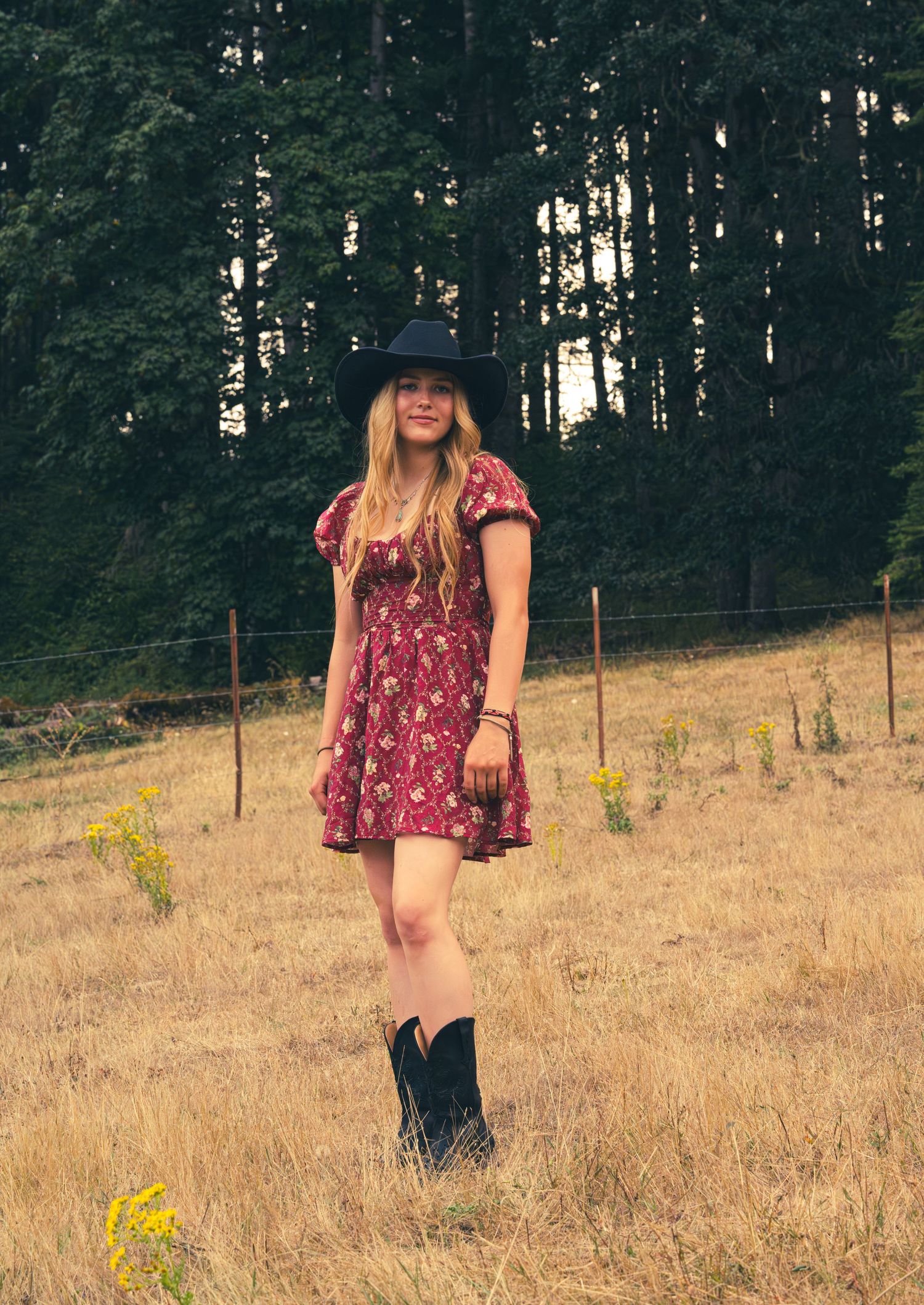 Young woman in floral mini dress, black wide-brim hat, and cowboy boots standing in a golden meadow.