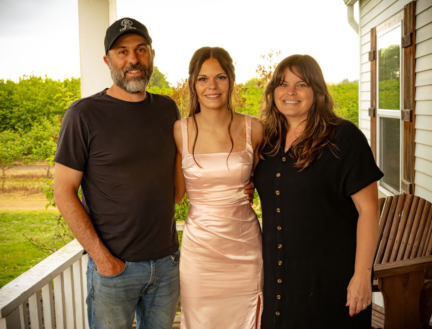 Family of three posing together on a porch, young woman in pink dress flanked by parents in black outfits.