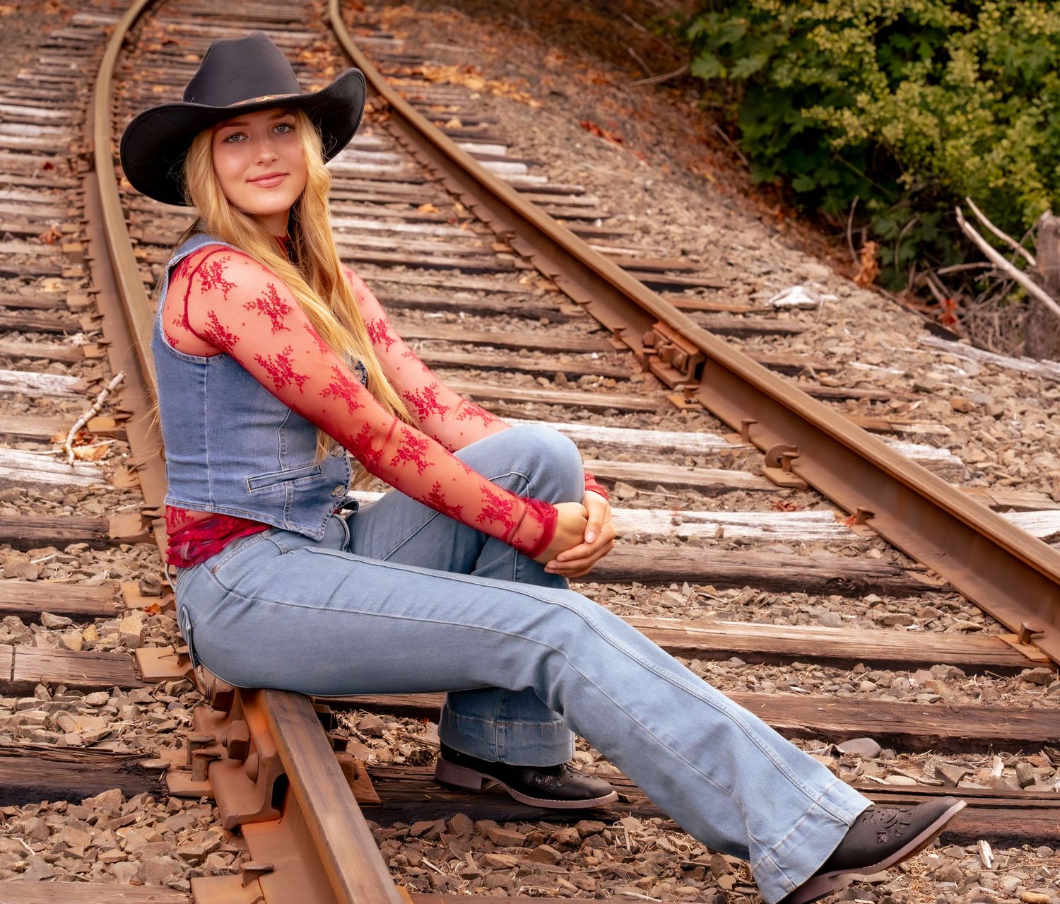 Young blonde woman in cowboy hat, jeans, and floral top sitting on railroad tracks outdoors.