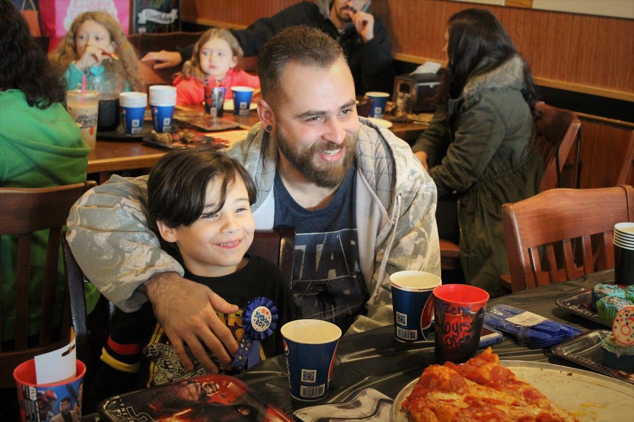 Smiling bearded man hugs young boy at birthday party table with pizza and drinks.