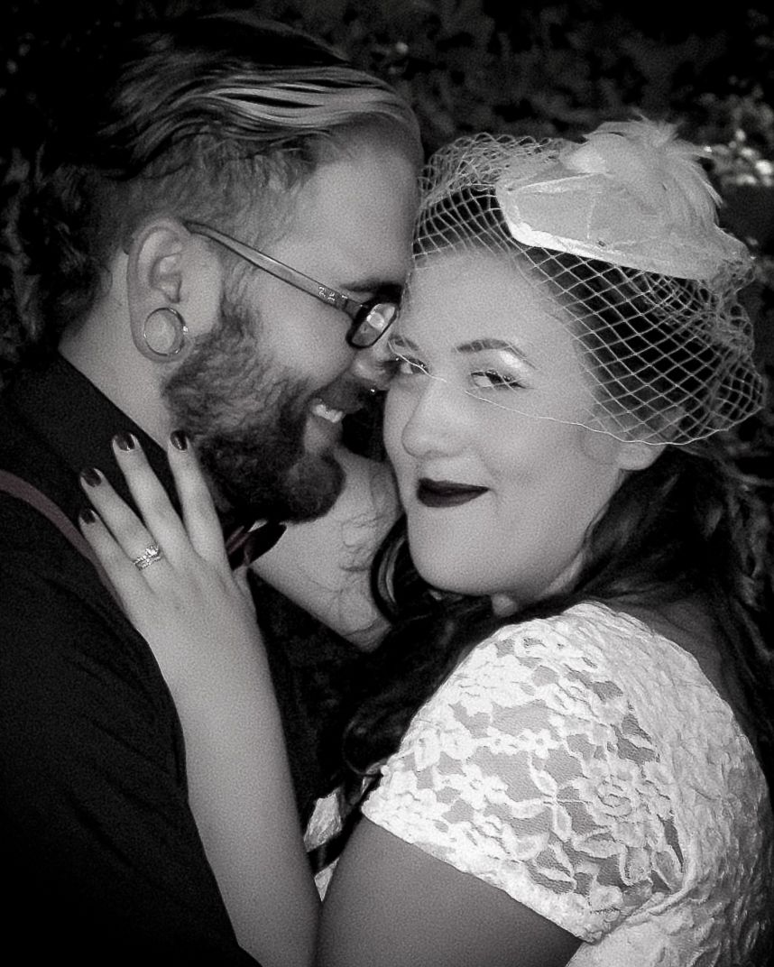 Black and white photo of a smiling bearded groom with glasses and bride wearing lace dress and birdcage veil touching foreheads.