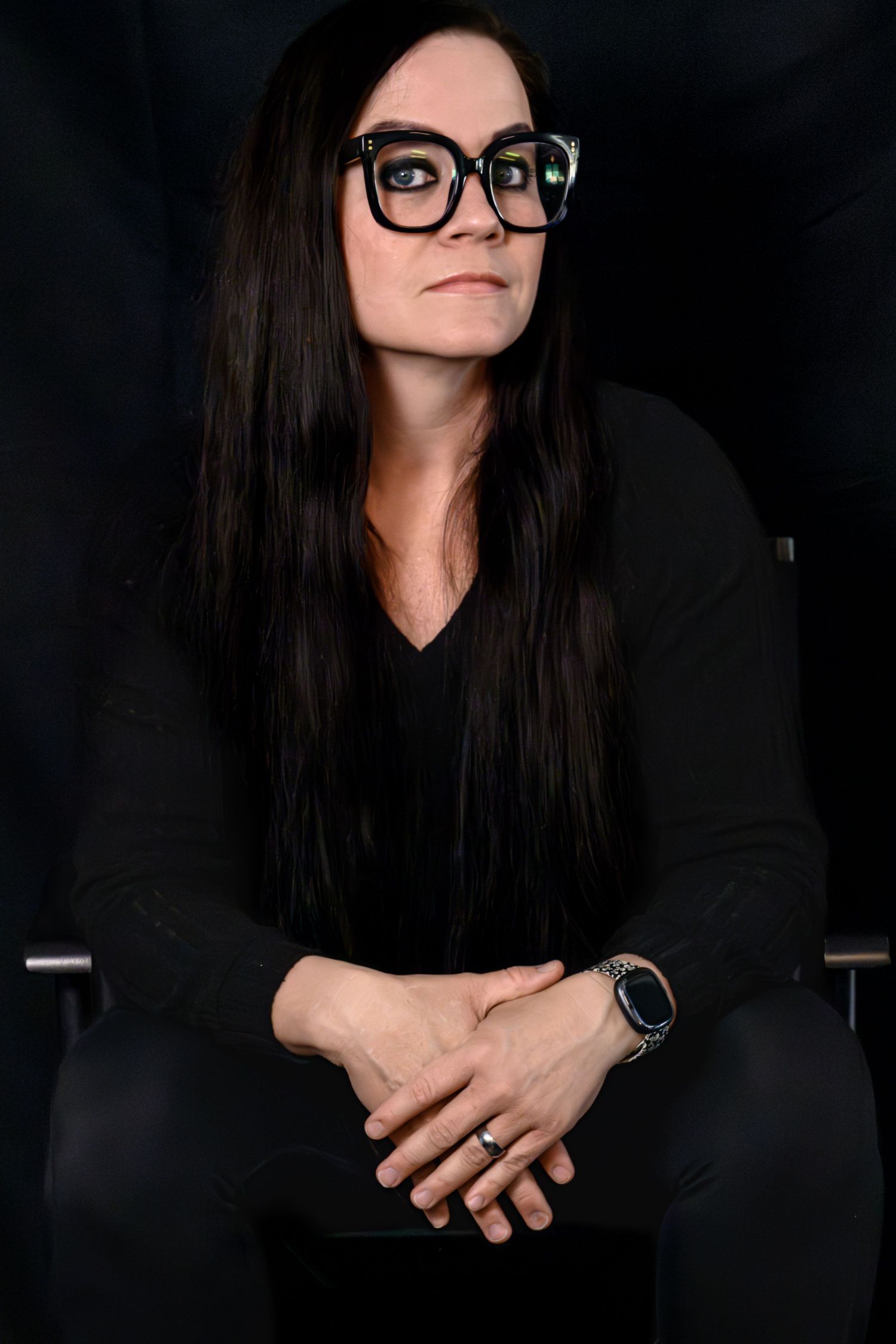 Woman with long dark hair and bold black glasses sits confidently in dark studio, wearing all black outfit.