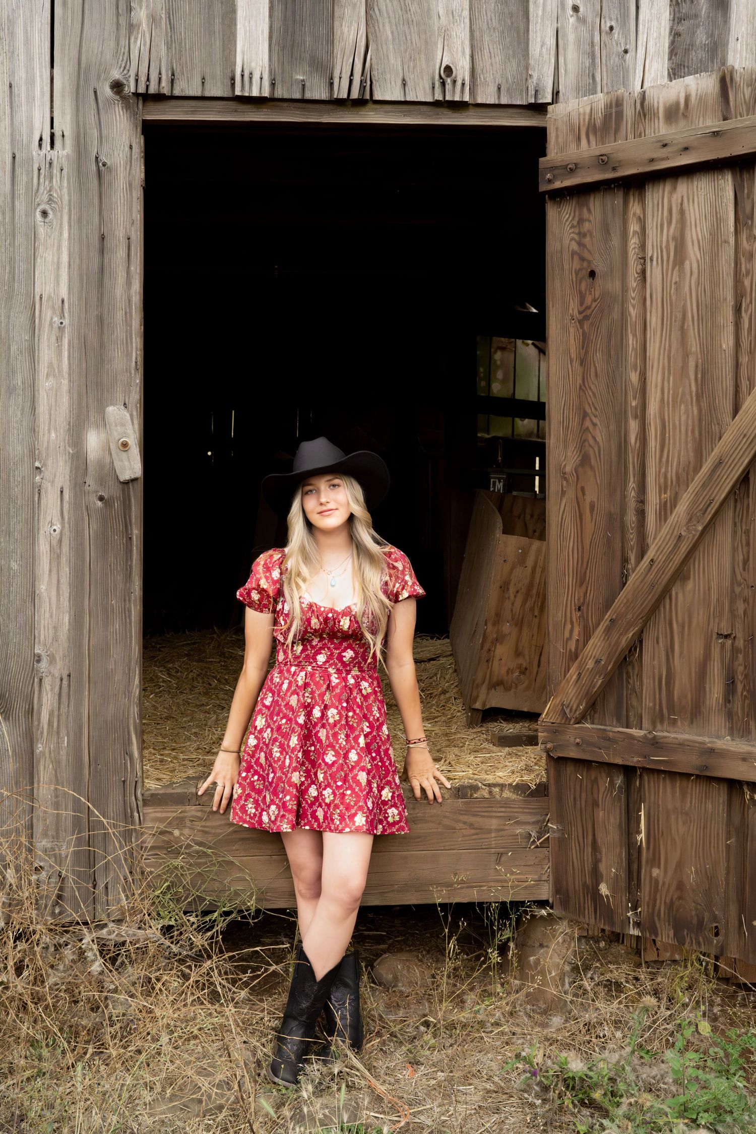 Blonde woman in red floral dress and black cowboy hat posing in rustic wooden barn doorway.