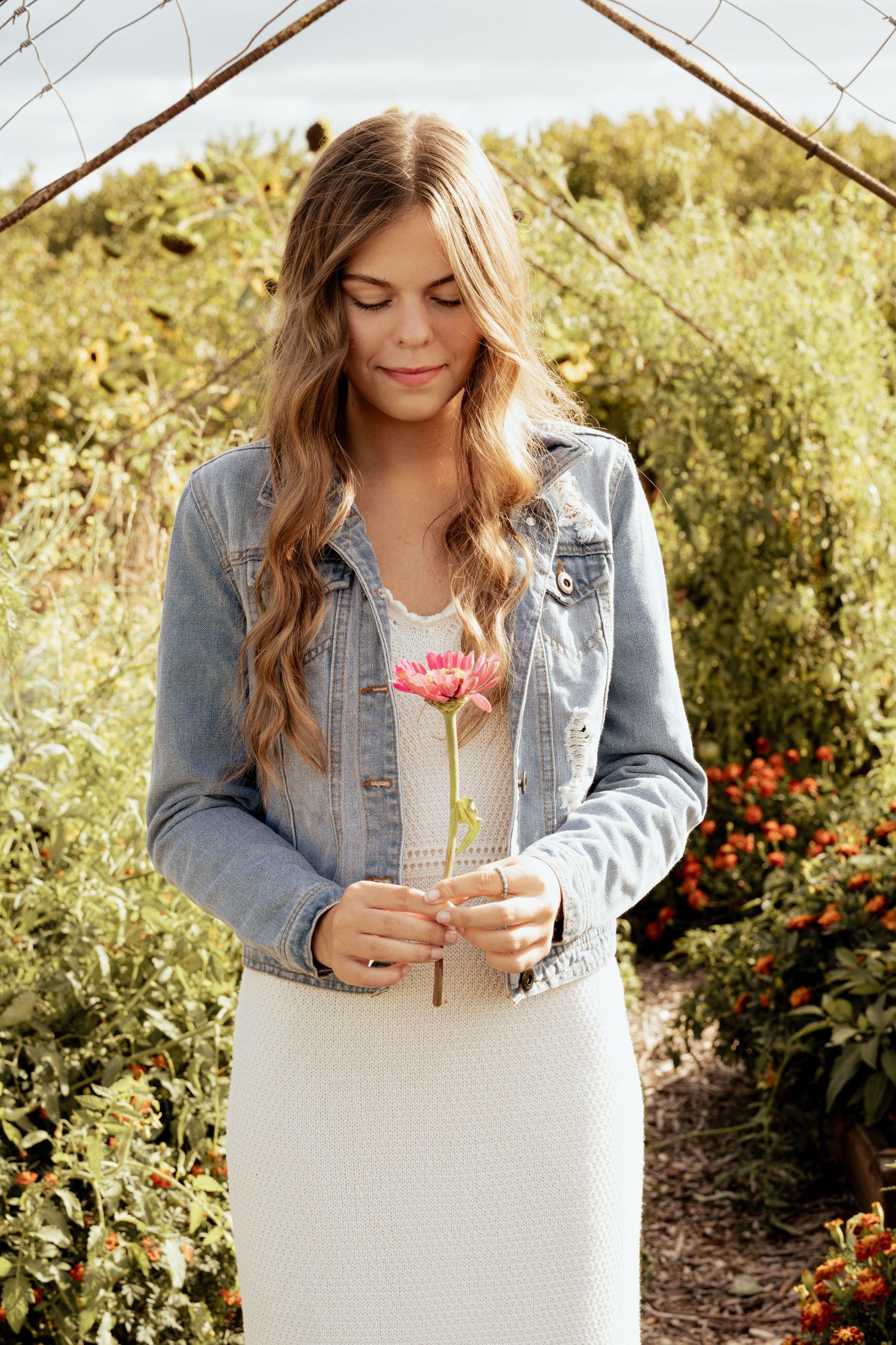 Young woman in denim jacket and white knit skirt holding a pink flower in a greenhouse.