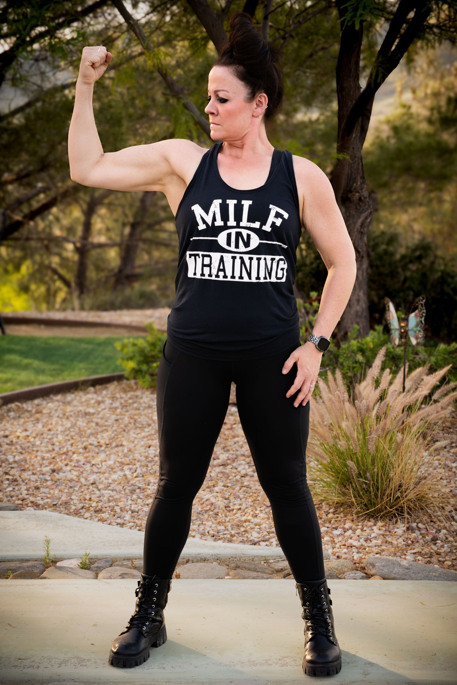 Woman in black MILF In Training tank top flexes bicep outdoors, wearing leggings and combat boots.