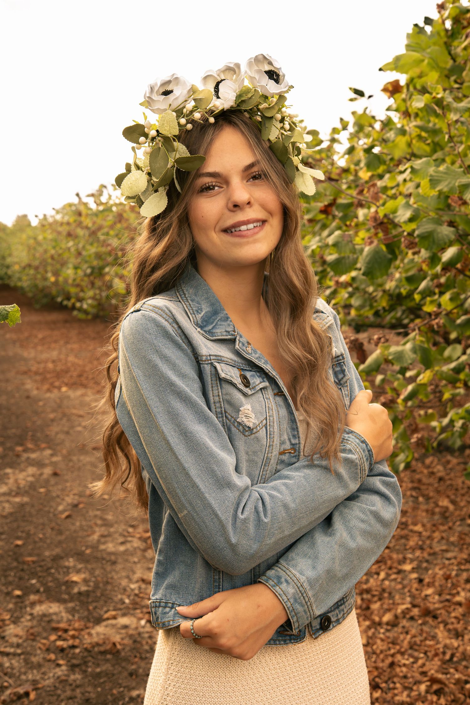 Smiling young woman wearing floral crown and denim jacket poses outdoors in lush green garden setting.