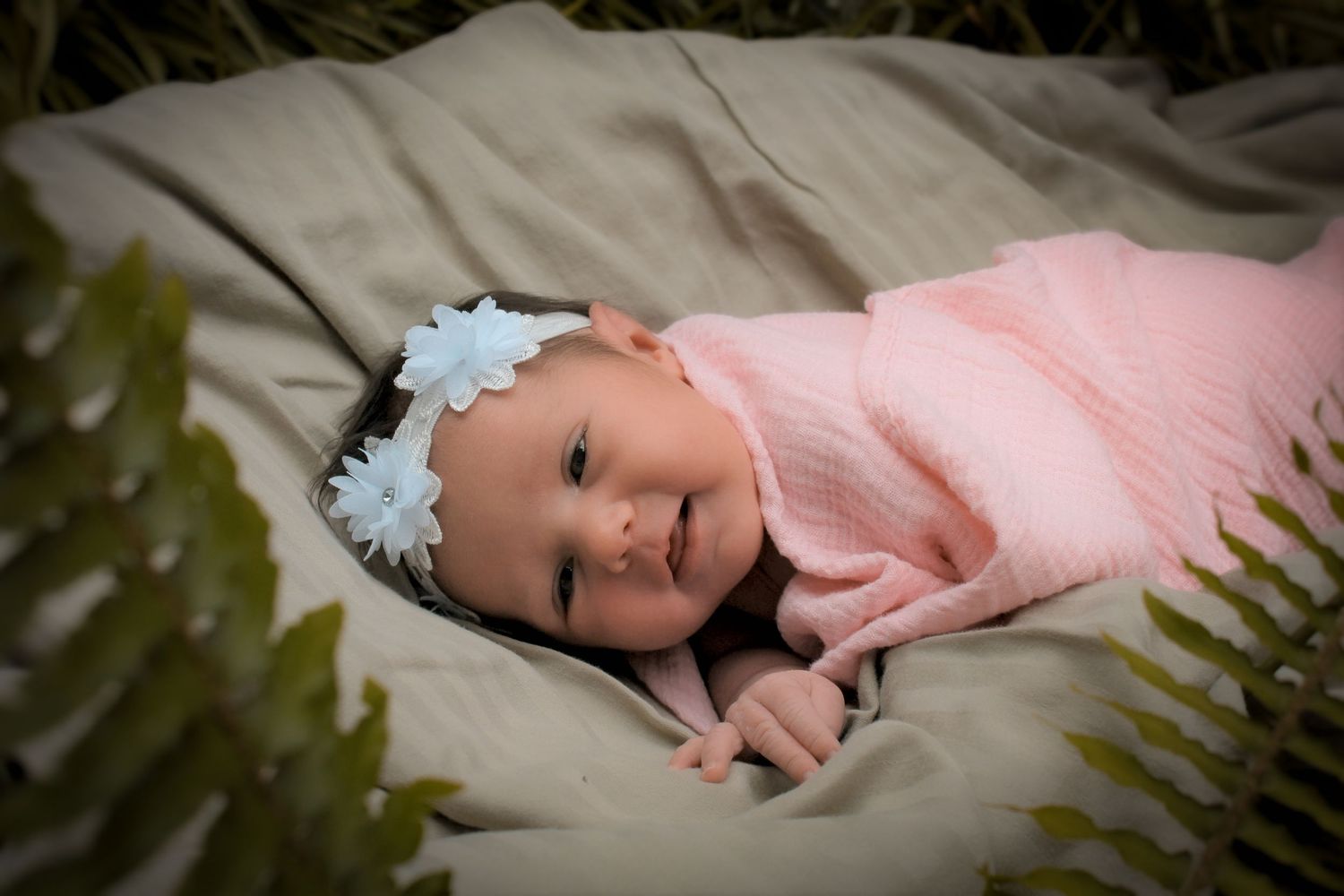 Smiling newborn baby in pink swaddle and white flower headband lying on blanket surrounded by ferns outdoors at Eve and Elm earthy Albany,OR